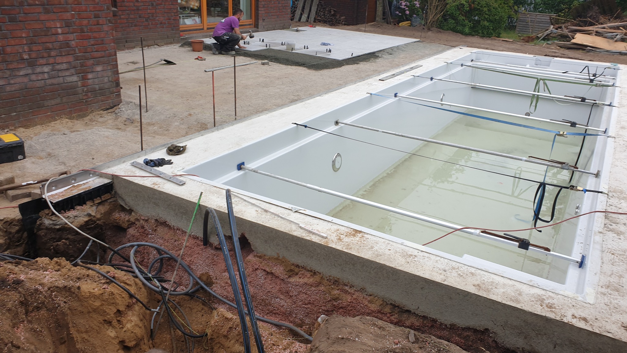 Budowa basenu: wykop, betonowa obudowa, instalacja. W tle pracownik układa płyty chodnikowe. Widoczne narzędzia i materiały budowlane.