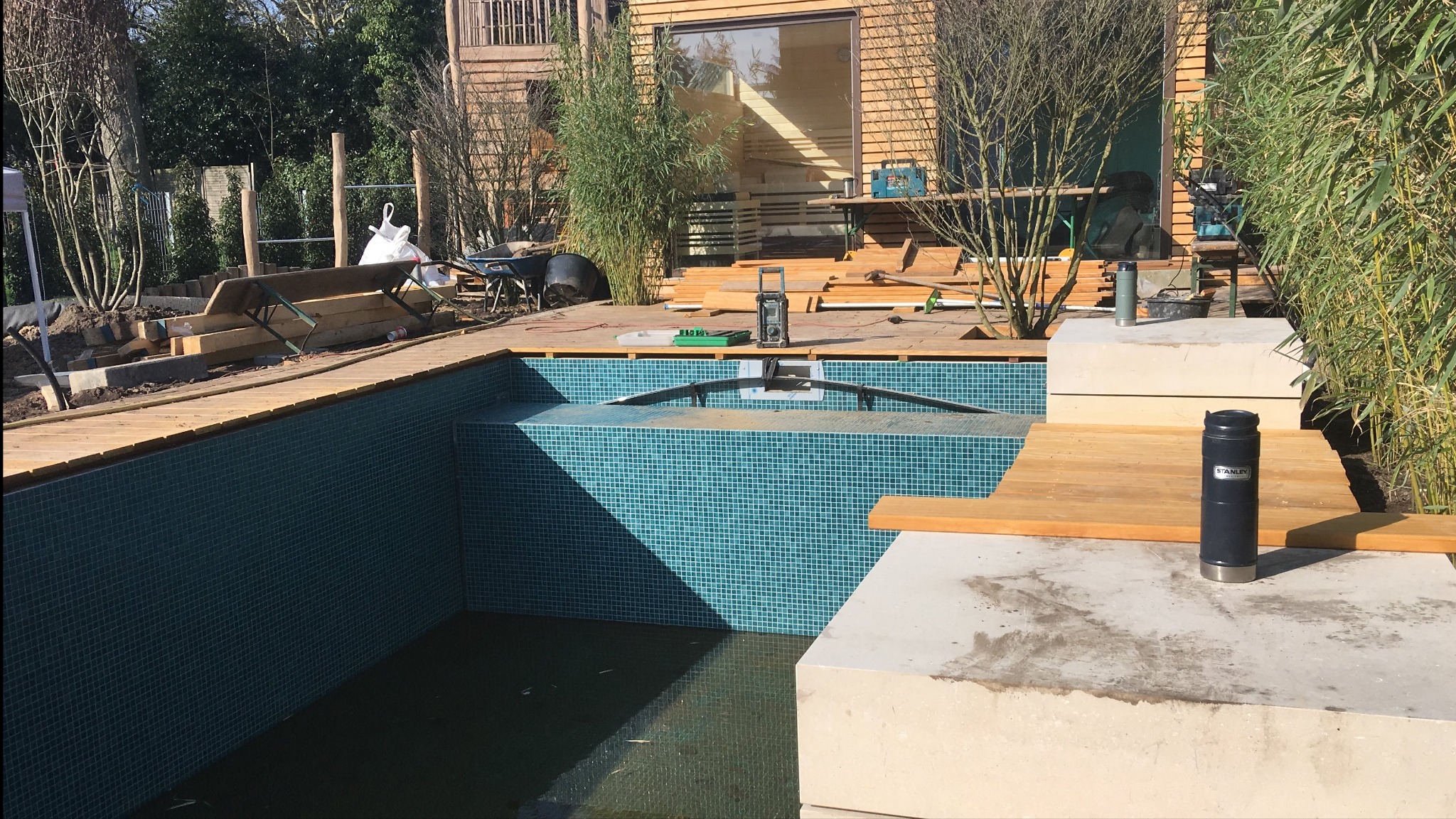 Budowa basenu z mozaiką i drewnianym tarasem w ogrodzie. Widoczne narzędzia i elementy konstrukcyjne. Termos Stanley na pierwszym planie.