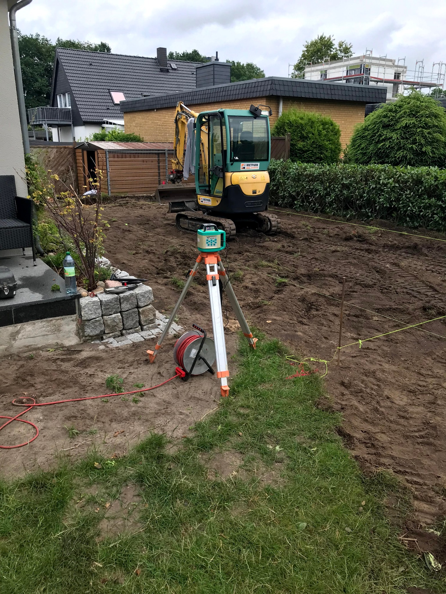 Mini koparka na placu budowy, niwelator laserowy na statywie, wytyczone linie. Przygotowanie terenu pod budowę, widoczne domy w tle.