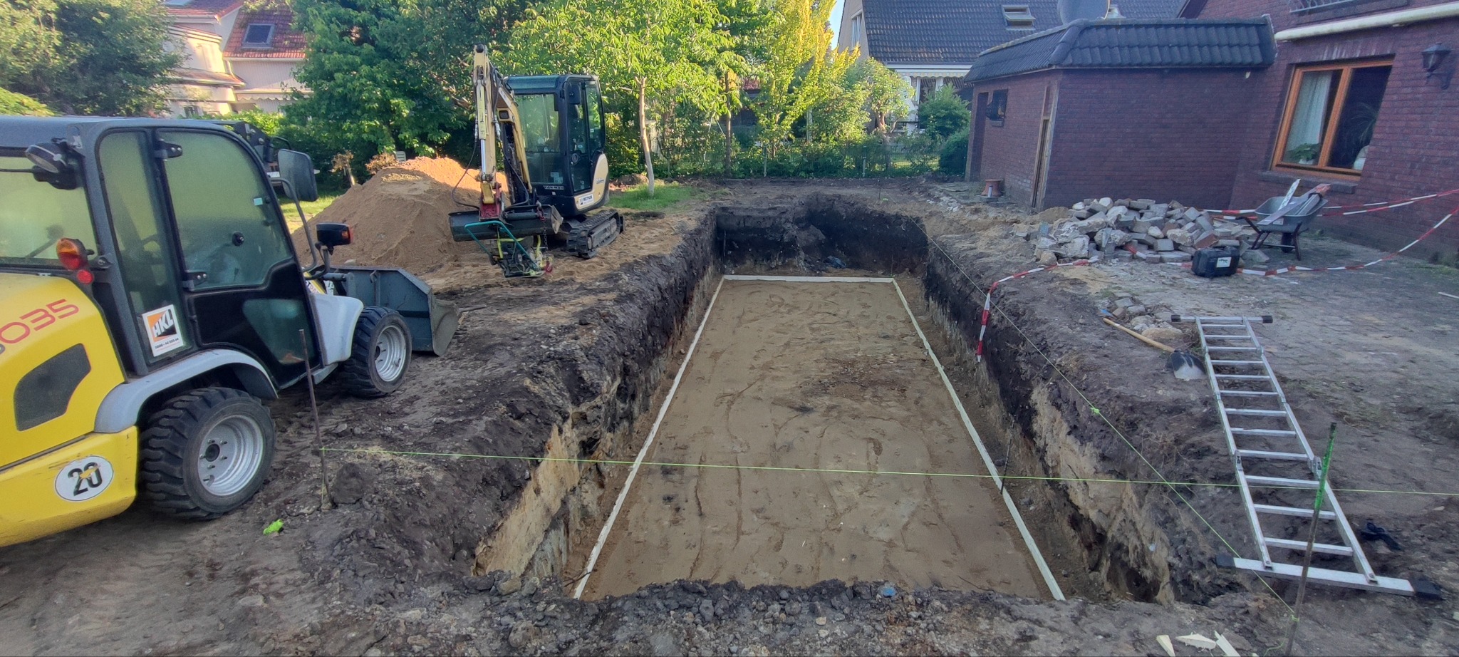 Wykop pod fundamenty z wytyczonym obrysem, widoczny sprzęt budowlany: minikoparka i ładowarka. Na drugim planie budynek mieszkalny i drzewa.