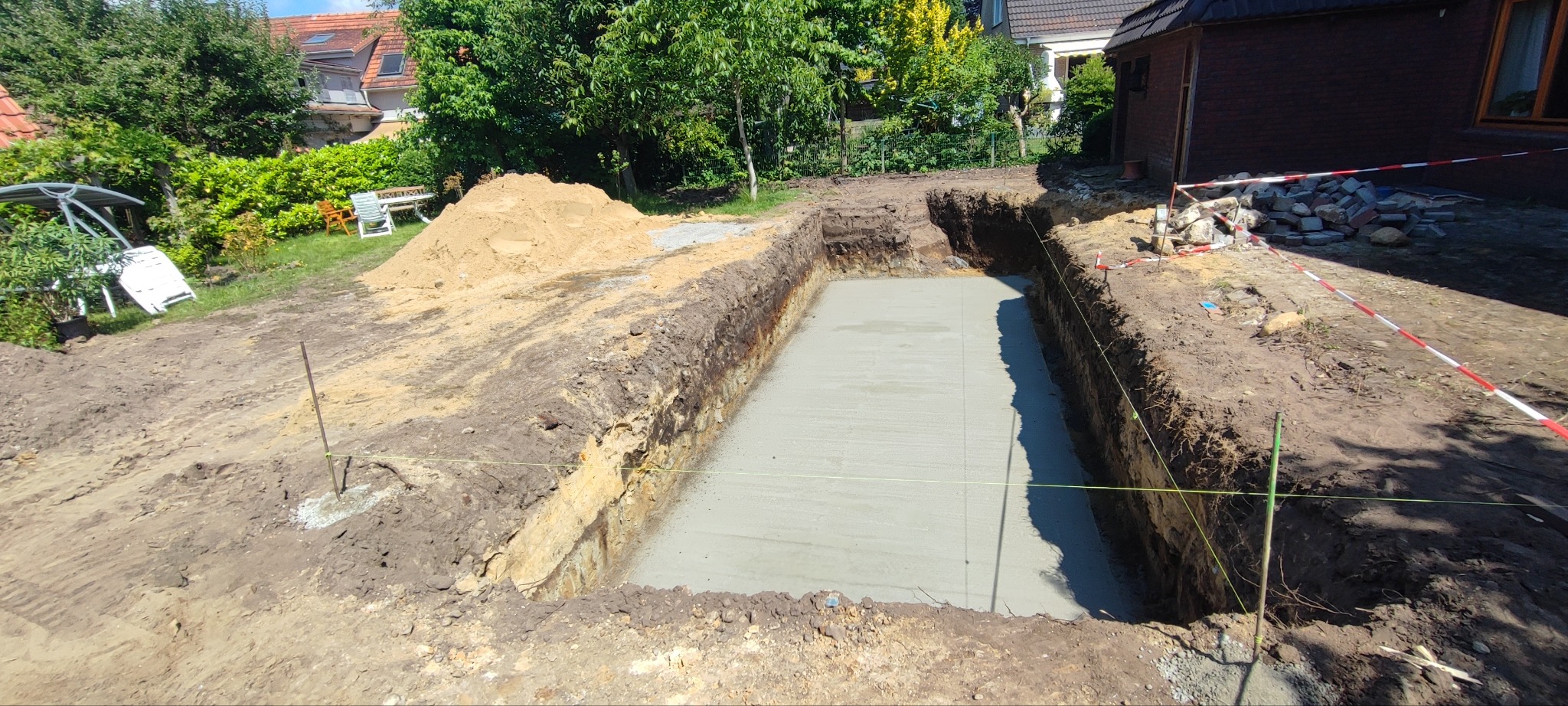 Wykop pod fundament z wylaną betonową płytą w ogrodzie. Widoczne zabezpieczenia taśmą ostrzegawczą i sterta ziemi. Prace ziemne w toku, przygotowanie pod budowę.