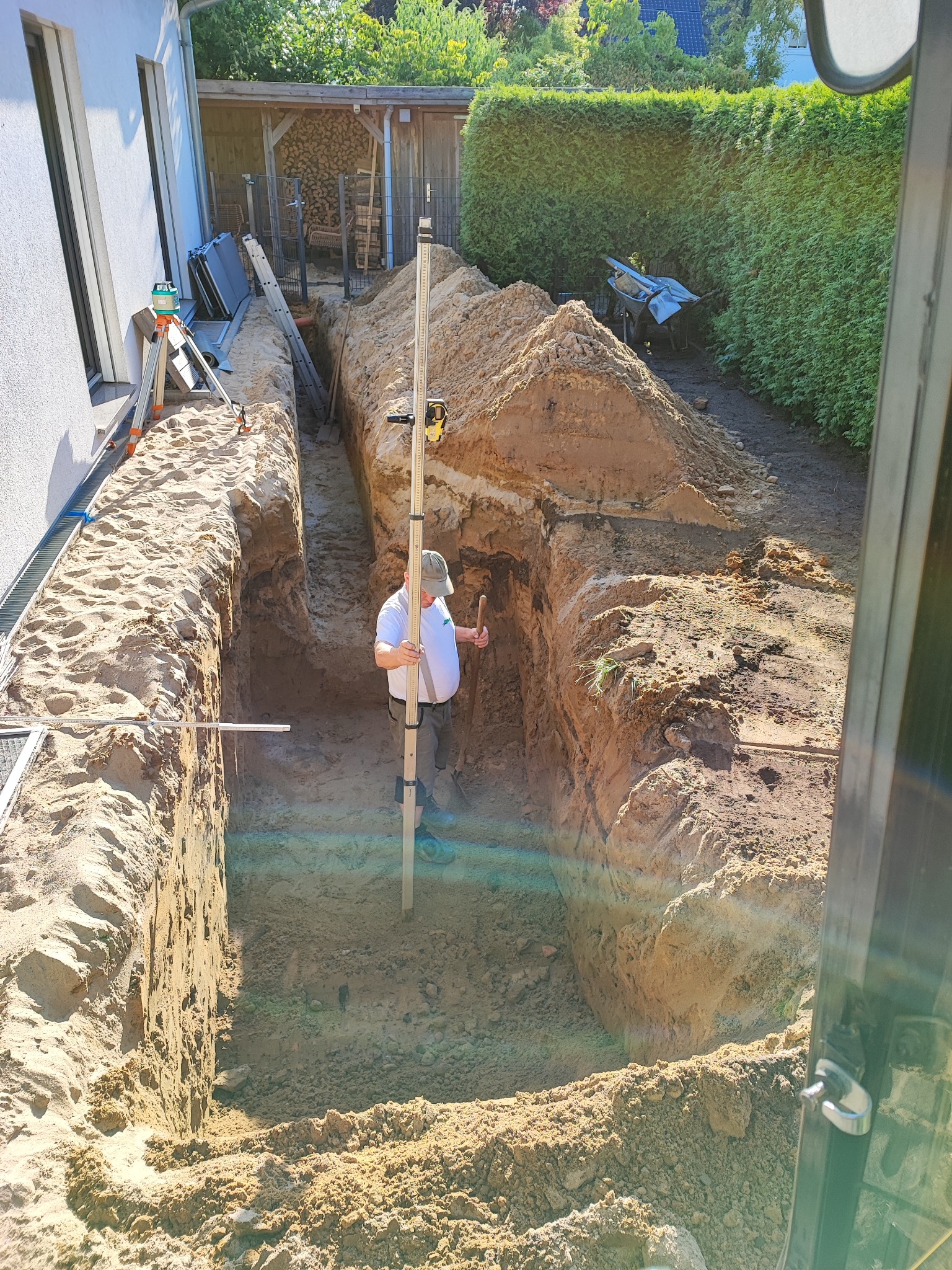 Wykop pod fundamenty domu jednorodzinnego, widok z kabiny koparki. Pracownik z łatą niwelacyjną kontroluje głębokość wykopu. W tle drewno na opał i zielony żywopłot.