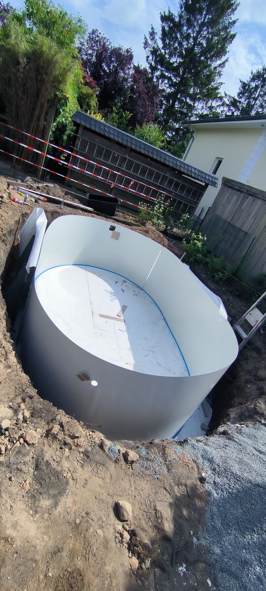 Montaż basenu ogrodowego: biała konstrukcja w wykopie, widoczne narzędzia i elementy przygotowawcze. Tło: zieleń ogrodu, dom. Ujęcie z góry, światło dzienne.