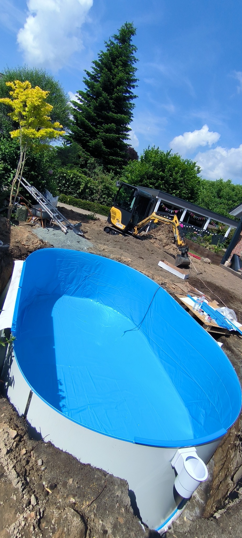 Montaż basenu ogrodowego z niebieską folią w wykopie, widoczny mały żółty koparka i zieleń ogrodu w tle, słoneczny dzień.