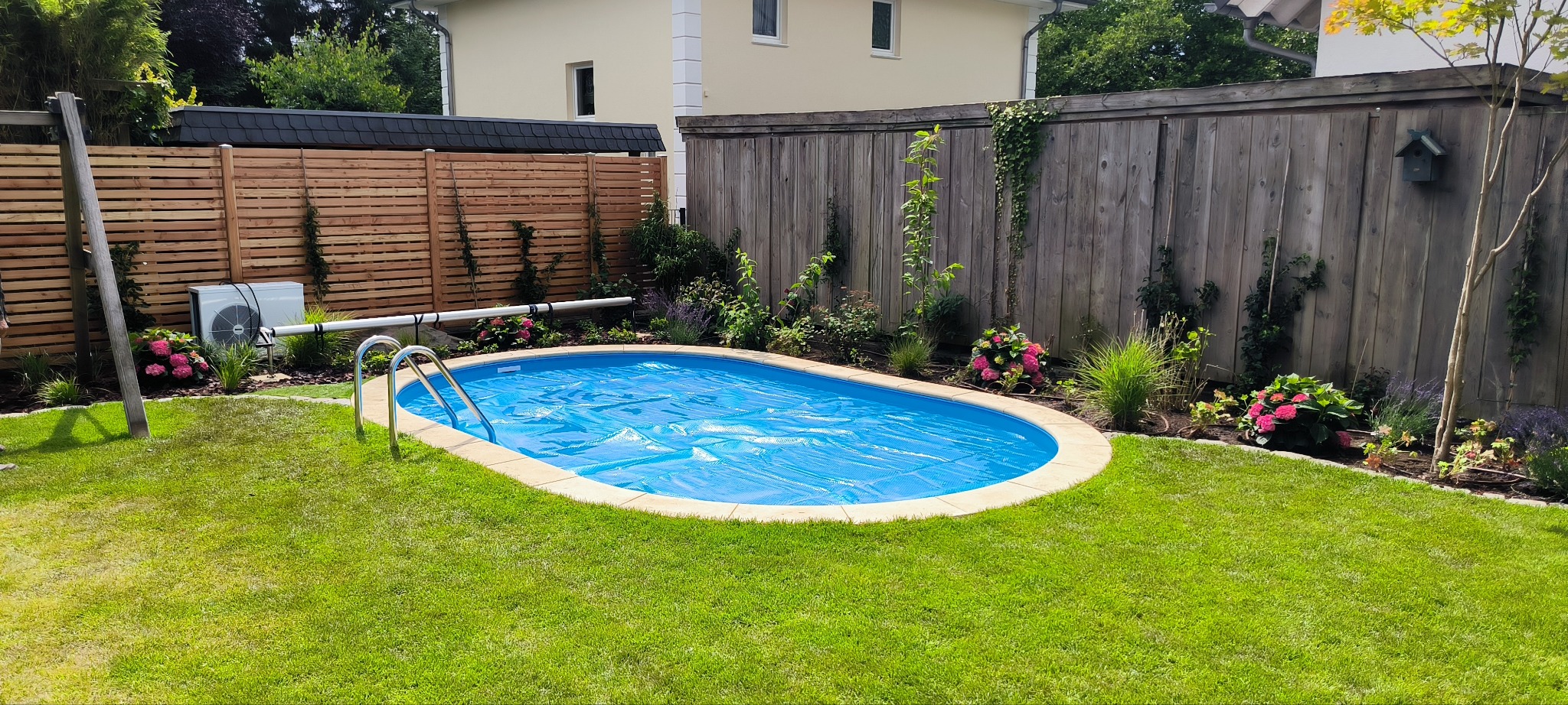 Owalny basen ogrodowy z niebieskim przykryciem, otoczony zielonym trawnikiem i rabatami kwiatowymi, na tle drewnianego ogrodzenia i domu. Słoneczny dzień.