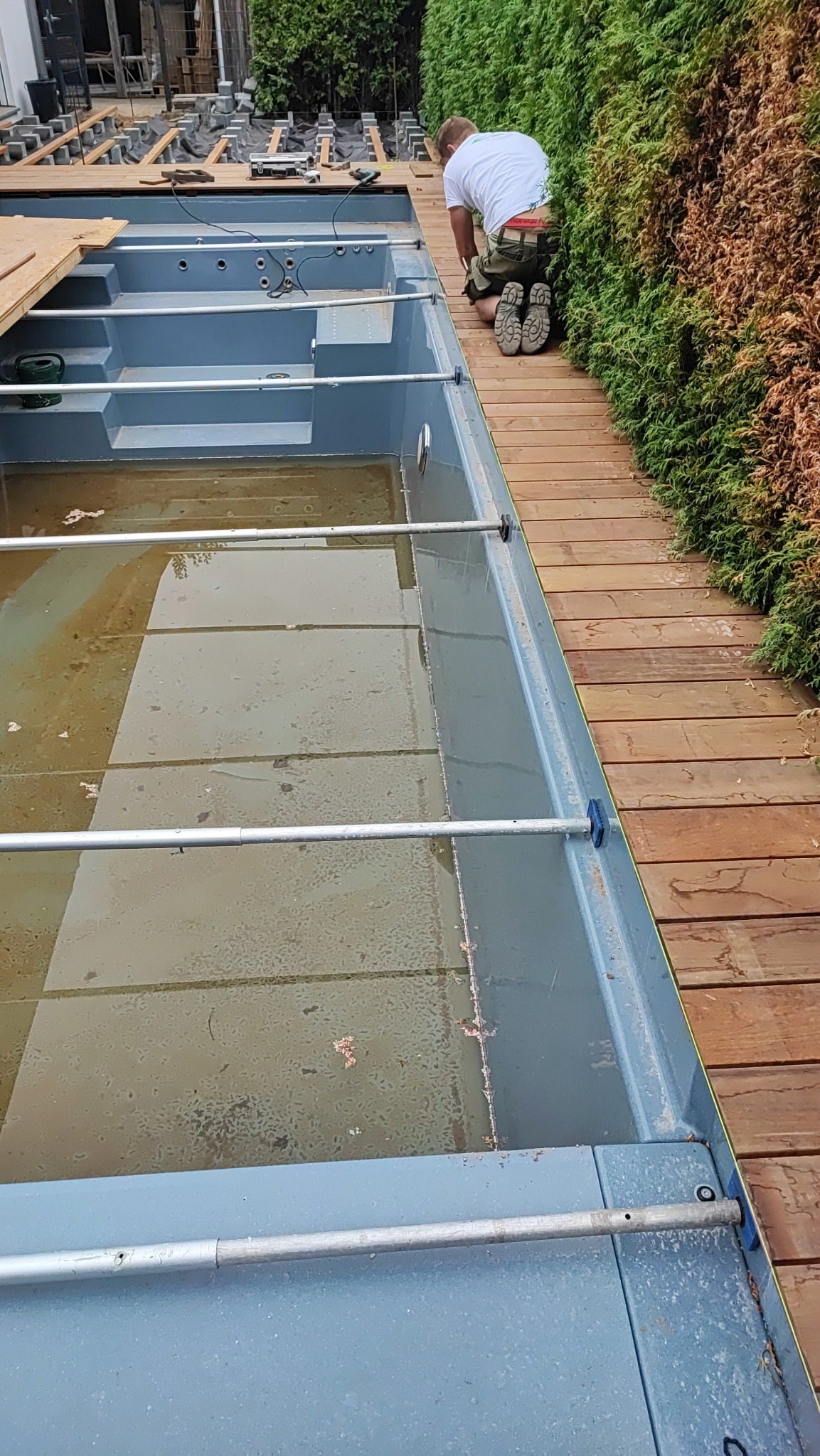 Montaż tarasu kompozytowego wokół basenu: pracownik na kolanach przy zielonym żywopłocie, układanie desek tarasowych. Widoczne elementy konstrukcyjne basenu i podbudowa tarasu.