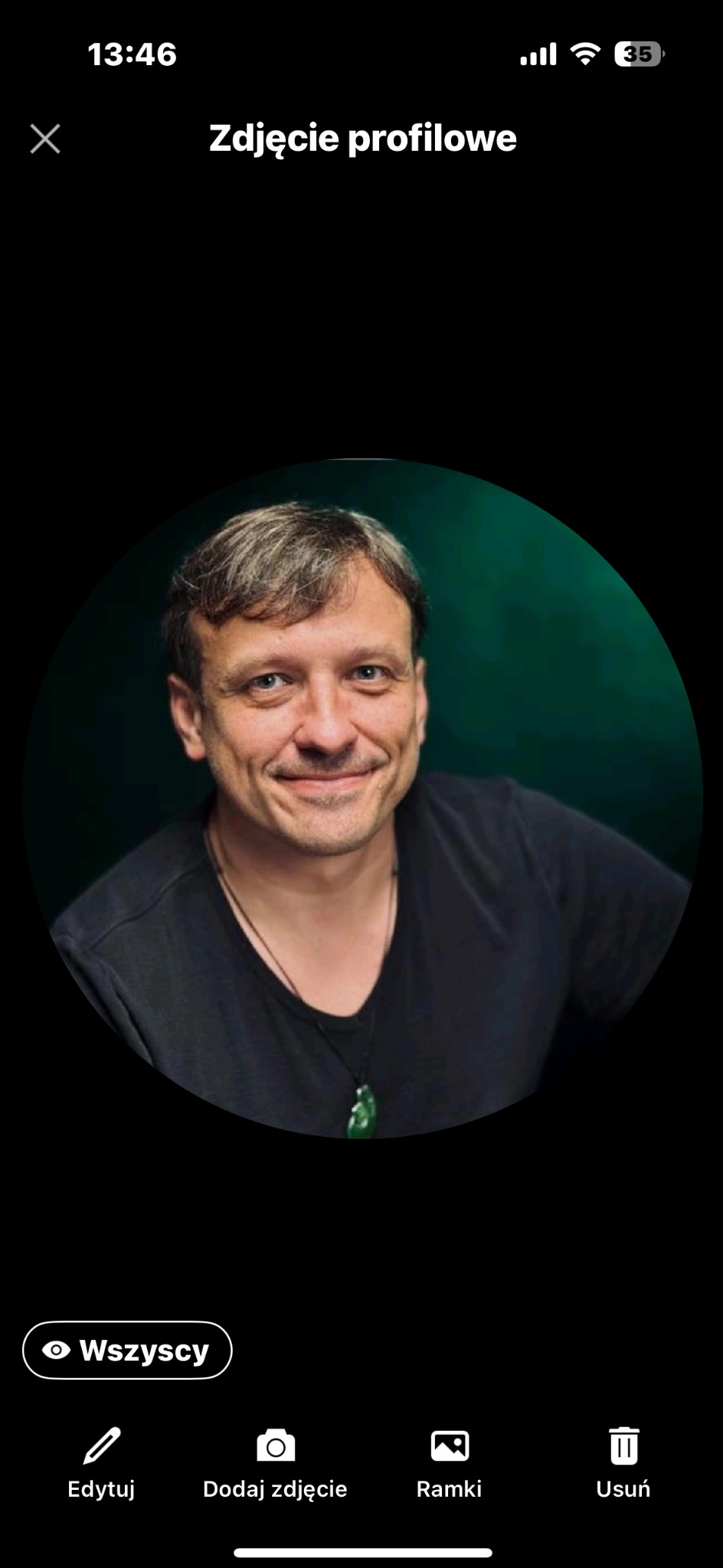 Portret uśmiechniętego mężczyzny z krótkimi włosami i niebieskimi oczami na ciemnozielonym tle. Ma na sobie czarną koszulkę i wisiorek.