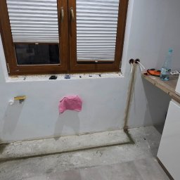 RALTECH USŁUGI ELEKTRYCZNE RADOSŁAW CZERNIAK - Wnętrze kuchni w trakcie remontu: widoczne wyprowadzone przewody elektryczne ze ściany, okno z żaluzjami, fragment zabudowy kuchennej i narzędzia na blacie.