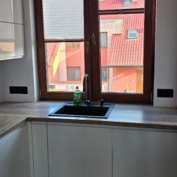 RALTECH USŁUGI ELEKTRYCZNE RADOSŁAW CZERNIAK - Nowoczesna kuchnia z białymi szafkami i czarnym zlewem pod oknem z widokiem na czerwone dachy budynków. Minimalistyczny design, czyste linie, neutralna kolorystyka.