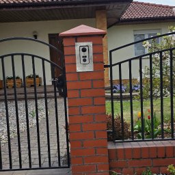 Elektryk - Domofon z kamerą zamontowany na ceglanym słupku ogrodzenia z metalową bramą, na tle domu z czerwoną dachówką i zielenią ogrodu.