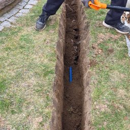 Elektryk - Wykop pod instalację elektryczną w Lipie Góry. Wąski rów w ziemi, widoczna niebieska rura karbowana, pracownicy z łopatami w tle. Prace ziemne.