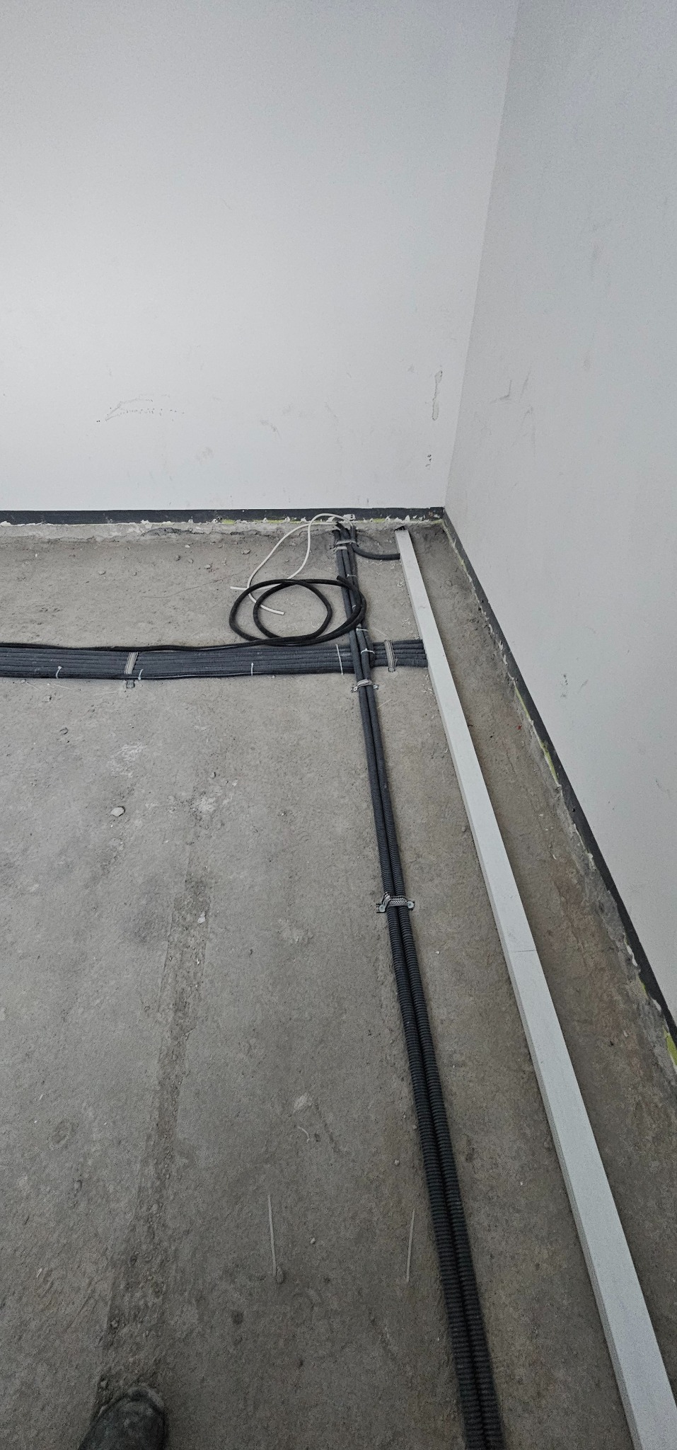 Szare rury instalacji elektrycznej ułożone na surowym betonie podłogi w rogu pomieszczenia, zabezpieczone metalowymi uchwytami, widoczne przewody.
