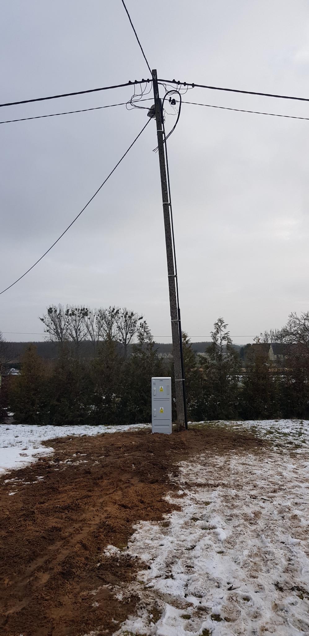 Słup elektryczny z zamontowaną skrzynką rozdzielczą na tle zimowego krajobrazu. Widoczne przewody, drabinka na słupie i częściowo ośnieżona ziemia.