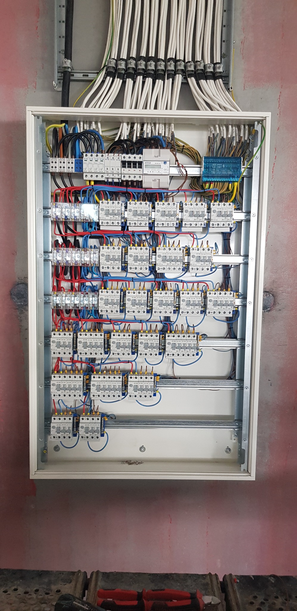 Rozdzielnica elektryczna z licznymi zabezpieczeniami i przewodami w kolorach: żółtym, niebieskim, czerwonym i czarnym. Nad rozdzielnicą wiązka kabli.