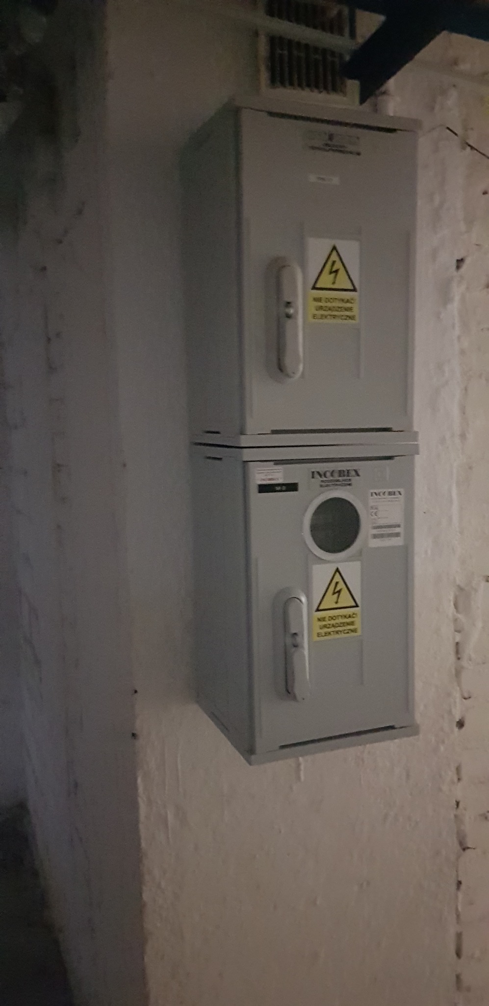 Dwa szare liczniki elektryczne IncoBex, jeden nad drugim, zamontowane na pomalowanej na biało ścianie z cegieł w piwnicy. Na licznikach naklejki ostrzegawcze.