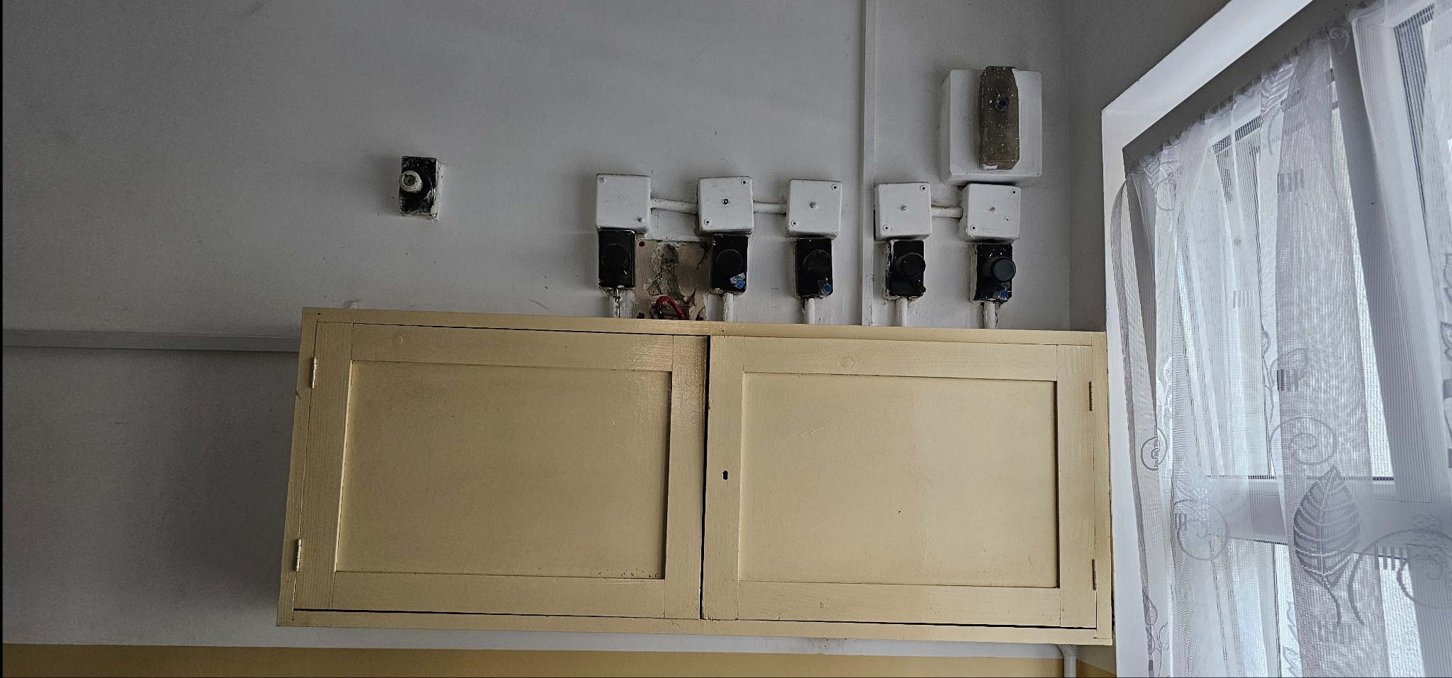 Szereg puszek elektrycznych z przewodami nad szafką w pomieszczeniu. Widoczne elementy instalacji elektrycznej i fragment okna z firanką.