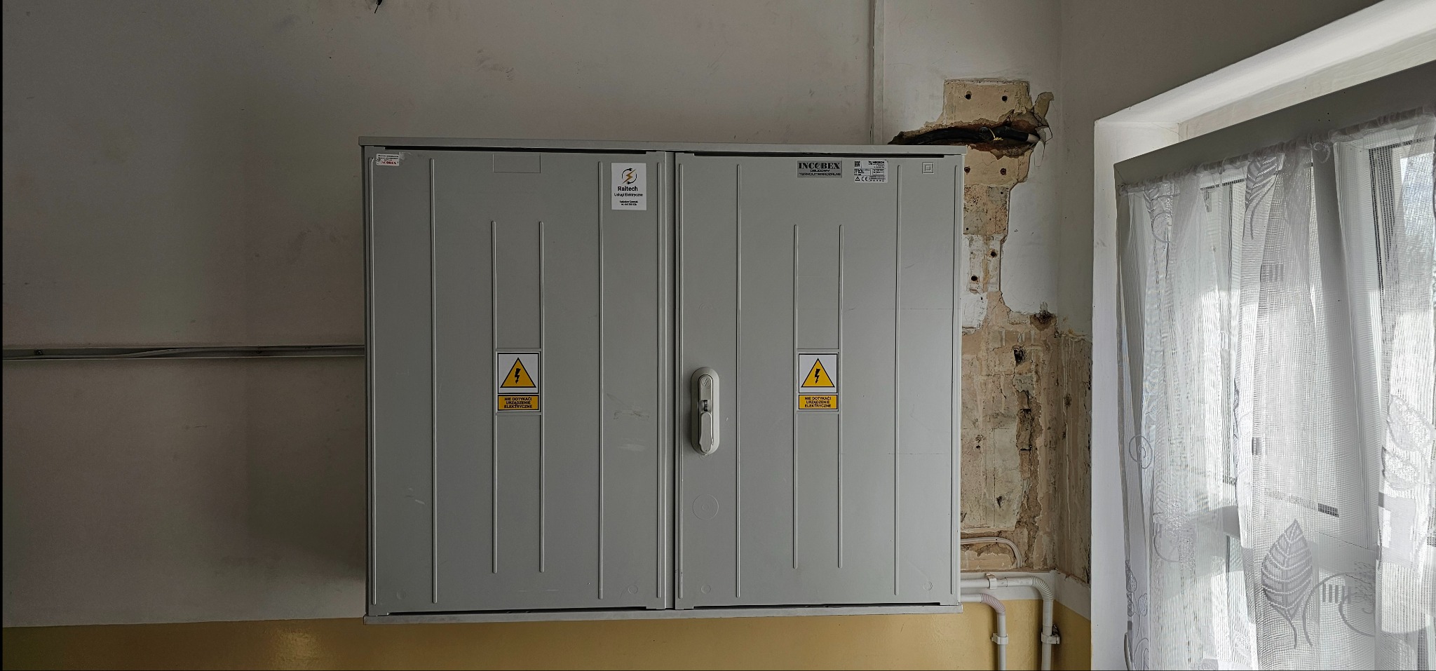 Szara, dwudrzwiowa skrzynka elektryczna z ostrzeżeniami, zamontowana na ścianie w pomieszczeniu z widocznymi uszkodzeniami tynku i oknem z firanką.