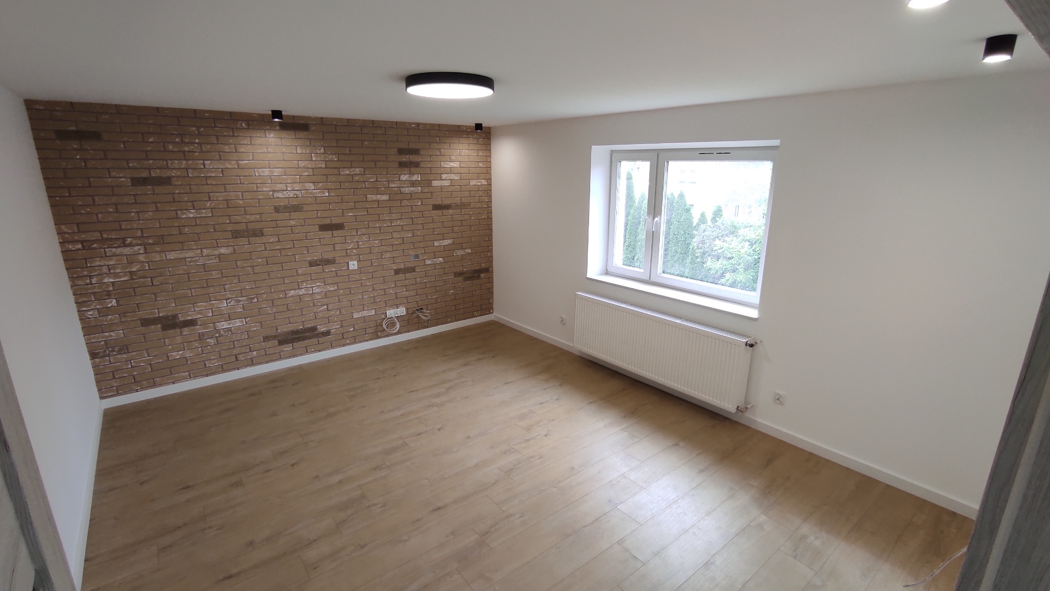 Jasne wnętrze po remoncie: drewnopodobna podłoga, ściana z cegły dekoracyjnej, białe ściany, okno z grzejnikiem. Minimalistyczne oświetlenie sufitowe.