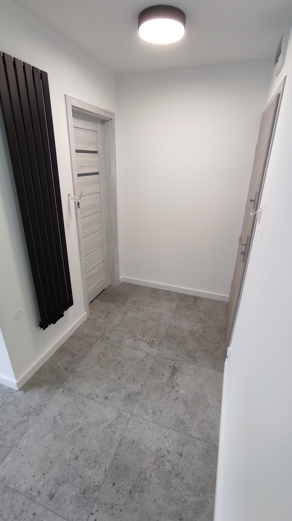 Nowoczesny przedpokój z szarą podłogą imitującą kamień, białe ściany, grzejnik panelowy i dwoje drzwi w odcieniach szarości. Minimalistyczny design i stonowana kolorystyka.