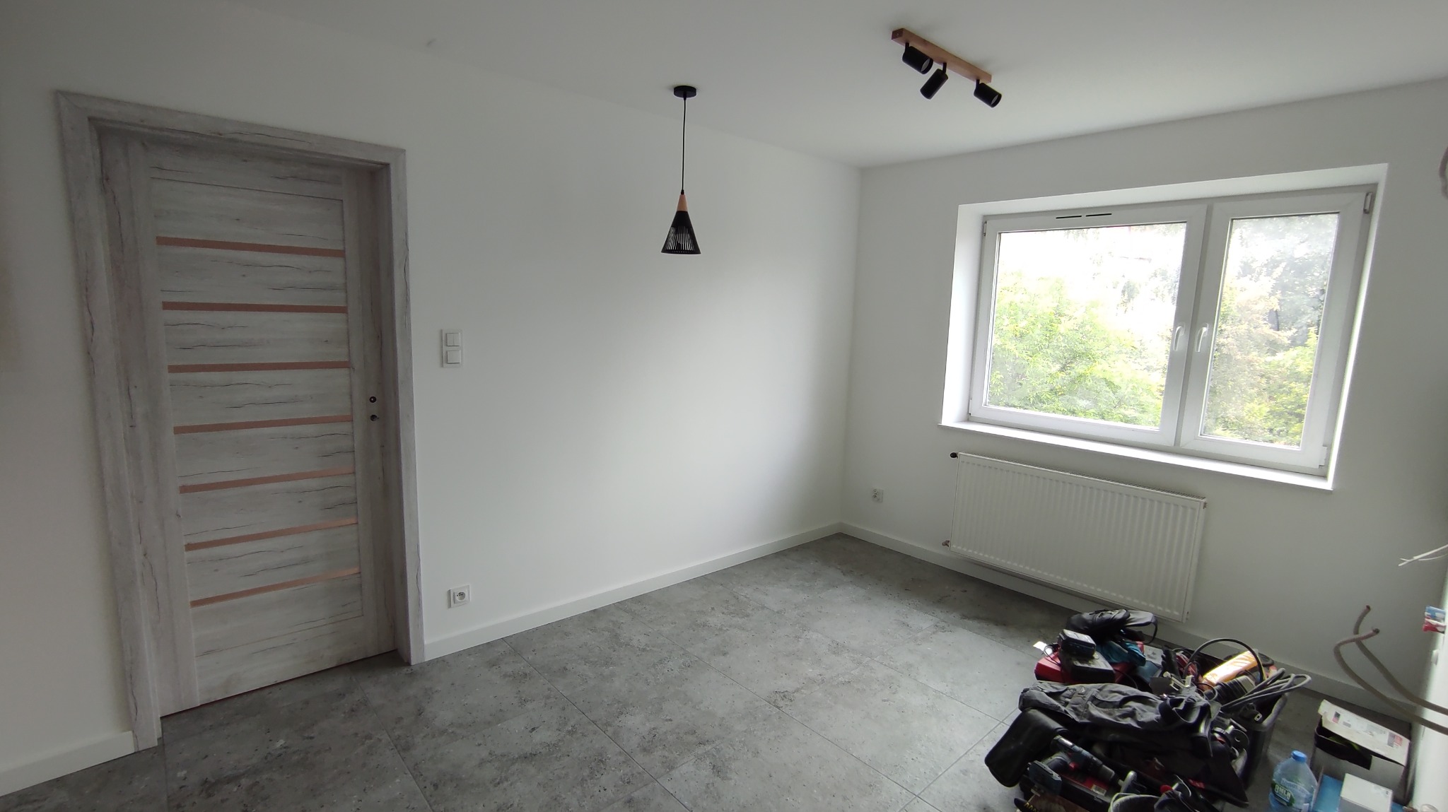 Jasne wnętrze pokoju po remoncie: białe ściany, szara podłoga, drewniane drzwi z poziomymi wstawkami, okno z widokiem na zieleń, lampa wisząca i reflektory sufitowe.