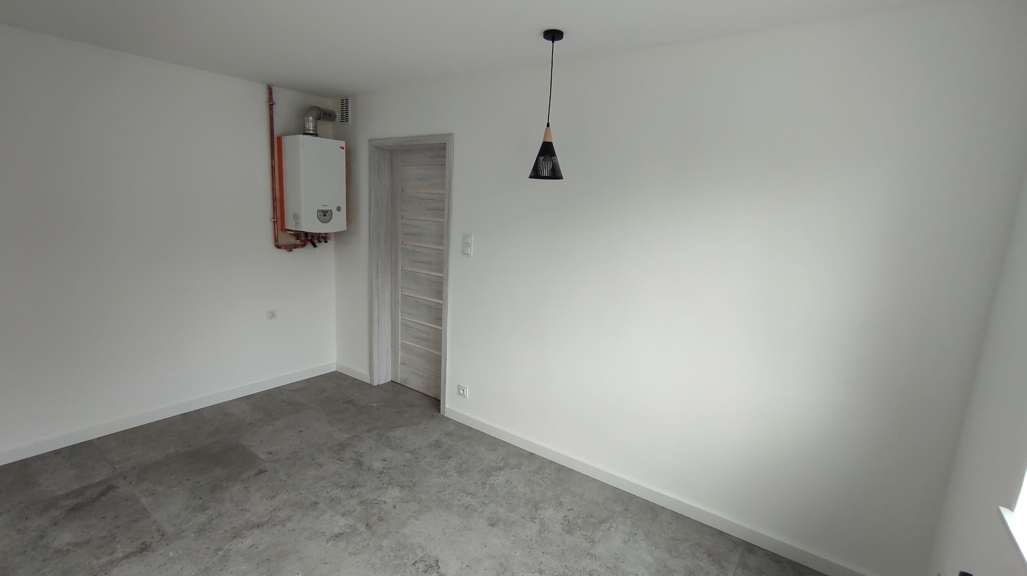 Wnętrze po remoncie w Szczawnie-Zdroju: białe ściany, szara podłoga, wiszący piec gazowy, drzwi z szarego drewna i czarna lampa wisząca. Minimalistyczny design.