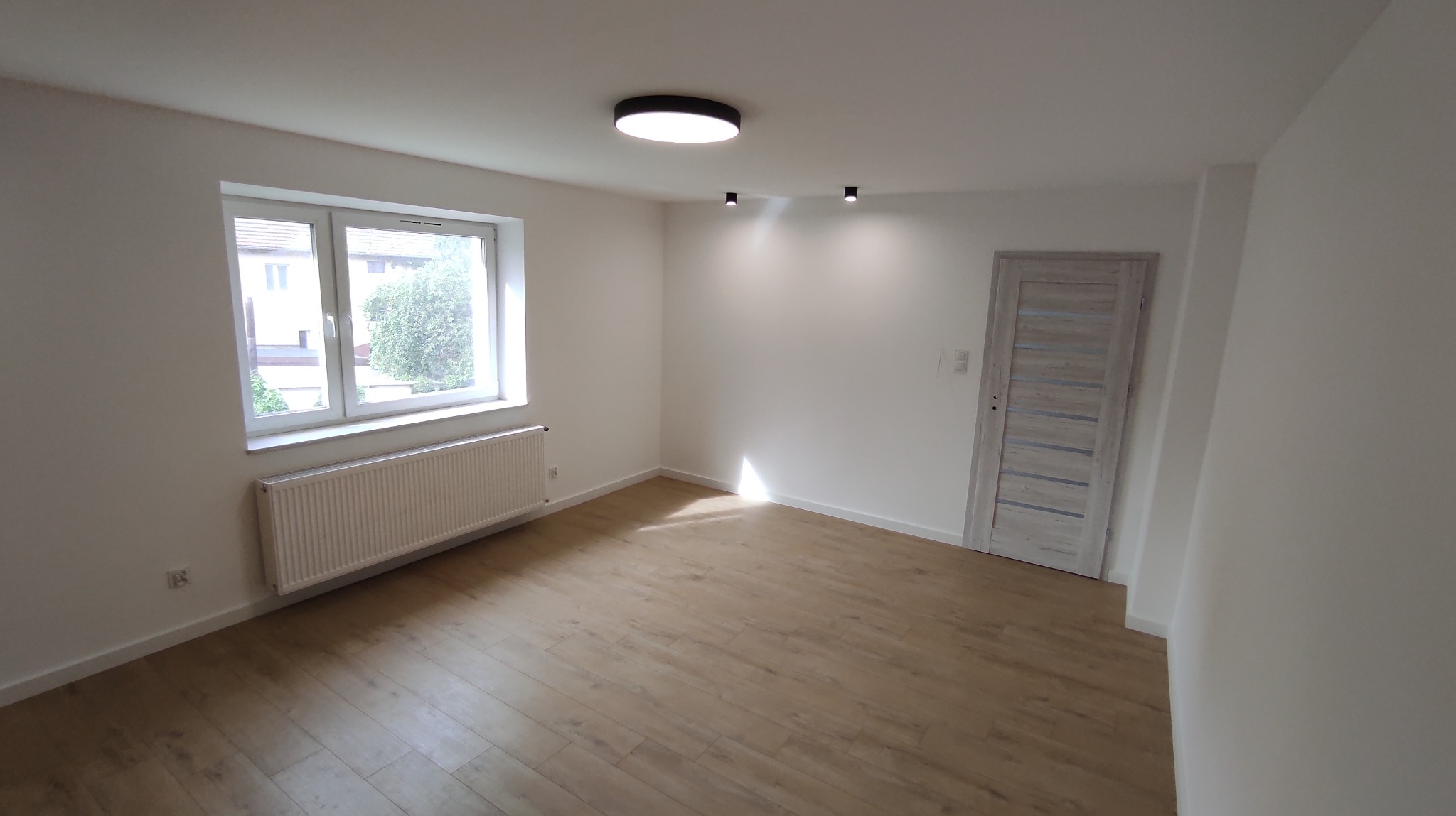 Jasne, puste wnętrze po remoncie: drewnopodobna podłoga, białe ściany, okno z grzejnikiem, drzwi z szarymi wstawkami i nowoczesne oświetlenie sufitowe.