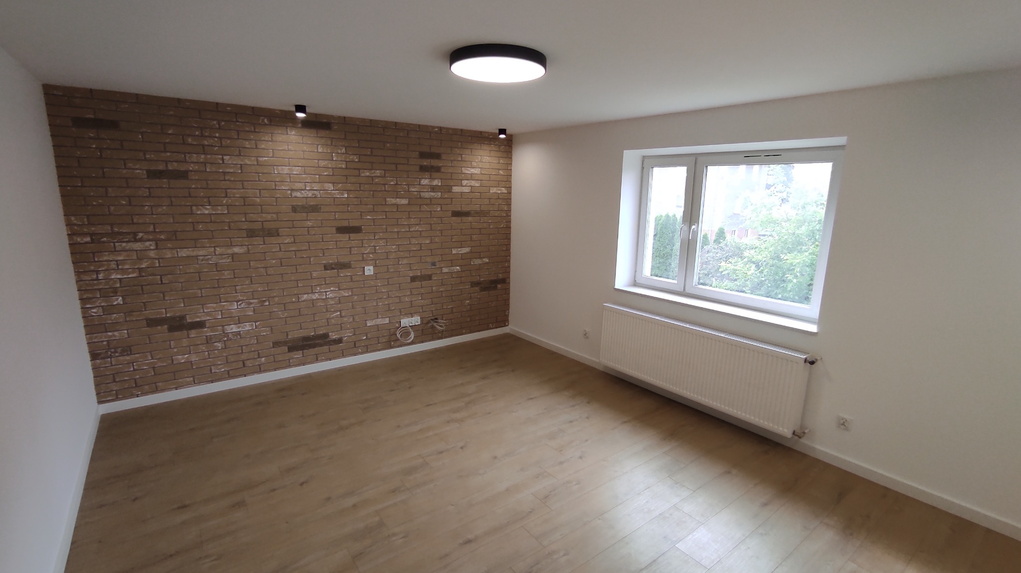 Jasne wnętrze po remoncie: parkiet, ściana z cegły dekoracyjnej, białe ściany, okno z widokiem na zieleń, grzejnik. Minimalistyczna lampa sufitowa.