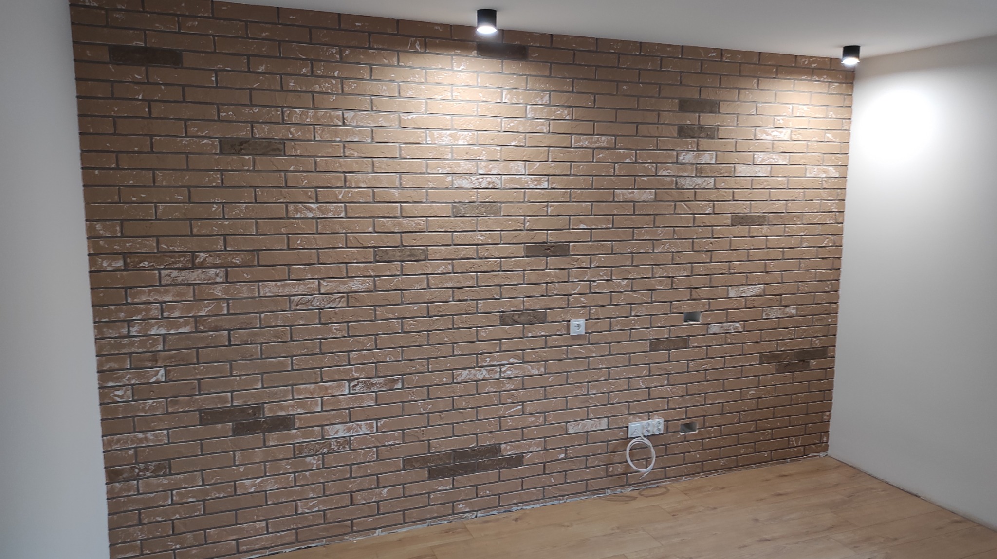 Ściana z cegły dekoracyjnej w odcieniach brązu, oświetlona punktowymi lampami sufitowymi. Widoczne gniazdka elektryczne i fragment drewnianej podłogi.