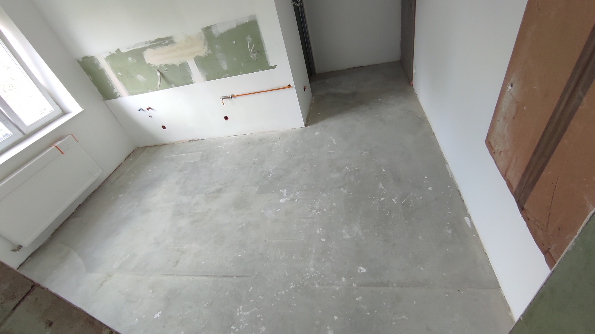 Wnętrze w trakcie remontu: szpachlowane ściany z widocznymi instalacjami, surowa podłoga, okno z grzejnikiem. Widok z góry, ukośny kadr.