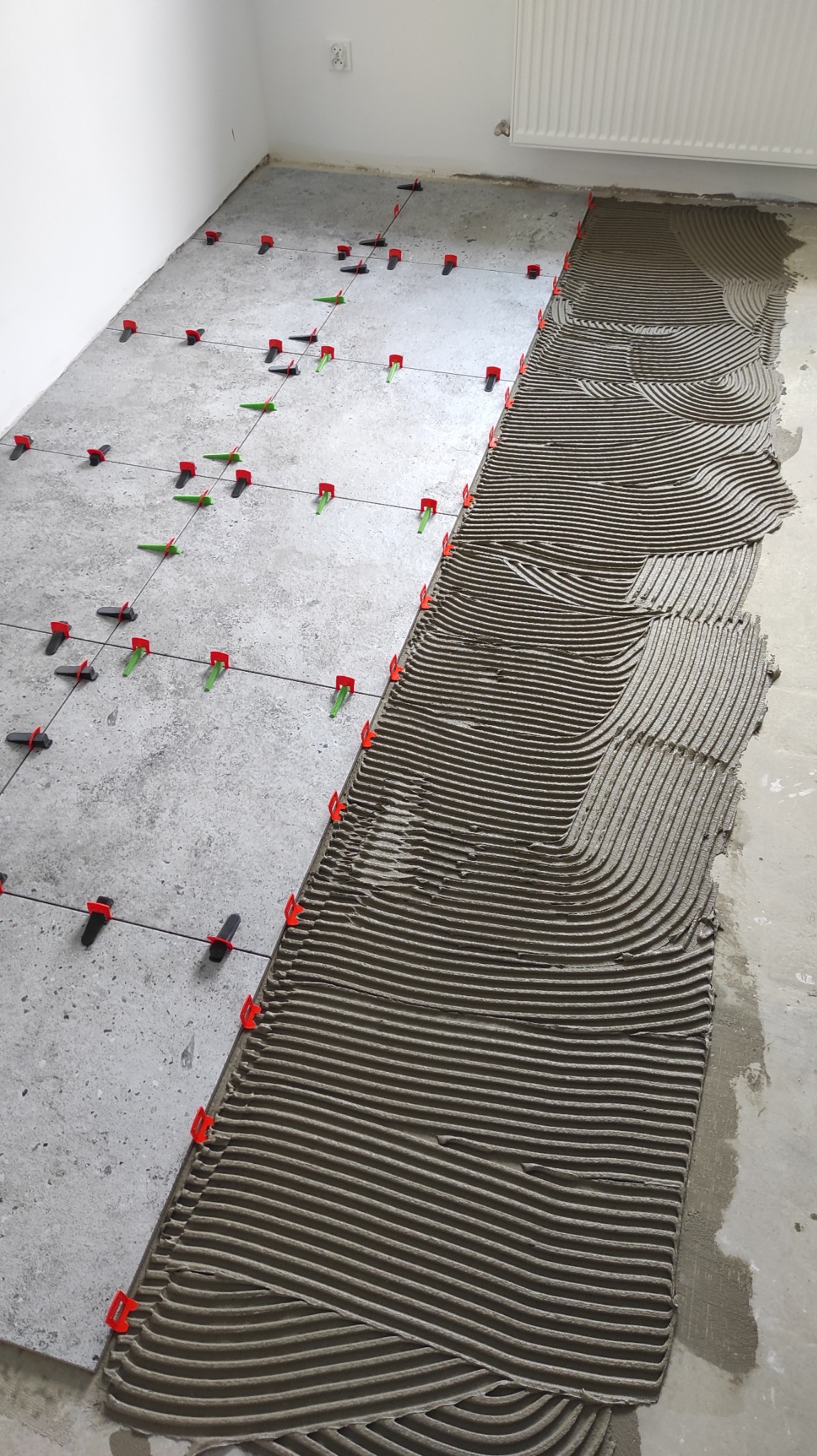 Układanie płytek podłogowych z użyciem systemu poziomowania: część płytek ułożona, część z nałożonym klejem w charakterystyczne prążki, widoczne klipsy i kliny.