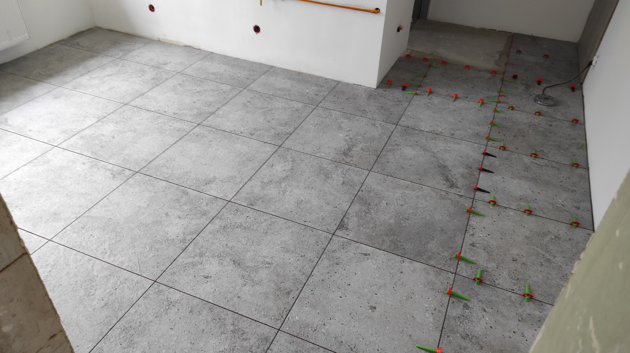 Ułożone szare płytki podłogowe z systemem poziomowania, widoczne czerwone i zielone kliny. Fragment ściany z instalacją elektryczną. Wnętrze w trakcie wykańczania.