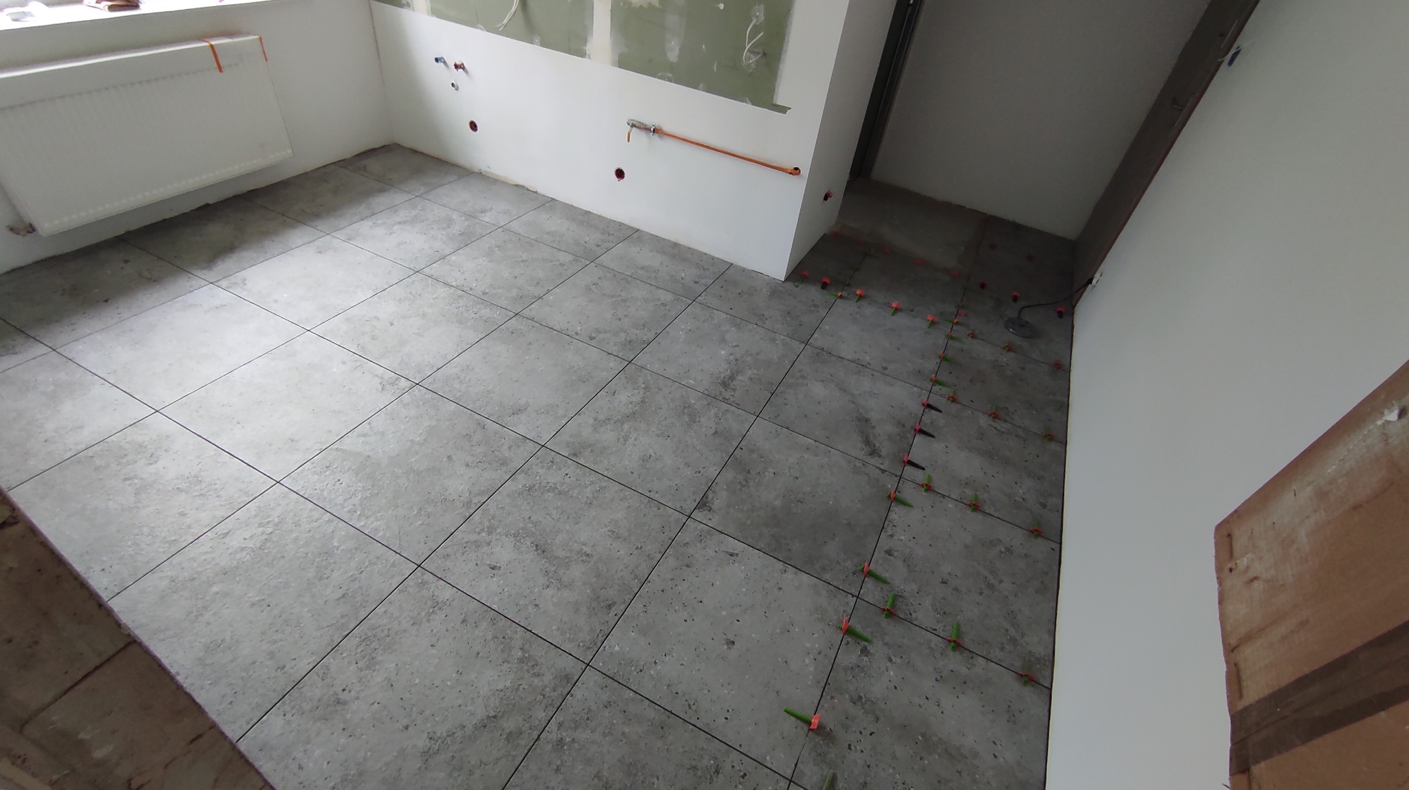 Ułożone szare płytki podłogowe w pomieszczeniu z białymi ścianami. Widoczne pomarańczowe i zielone kliny poziomujące. Częściowo wykończona ściana z instalacją.