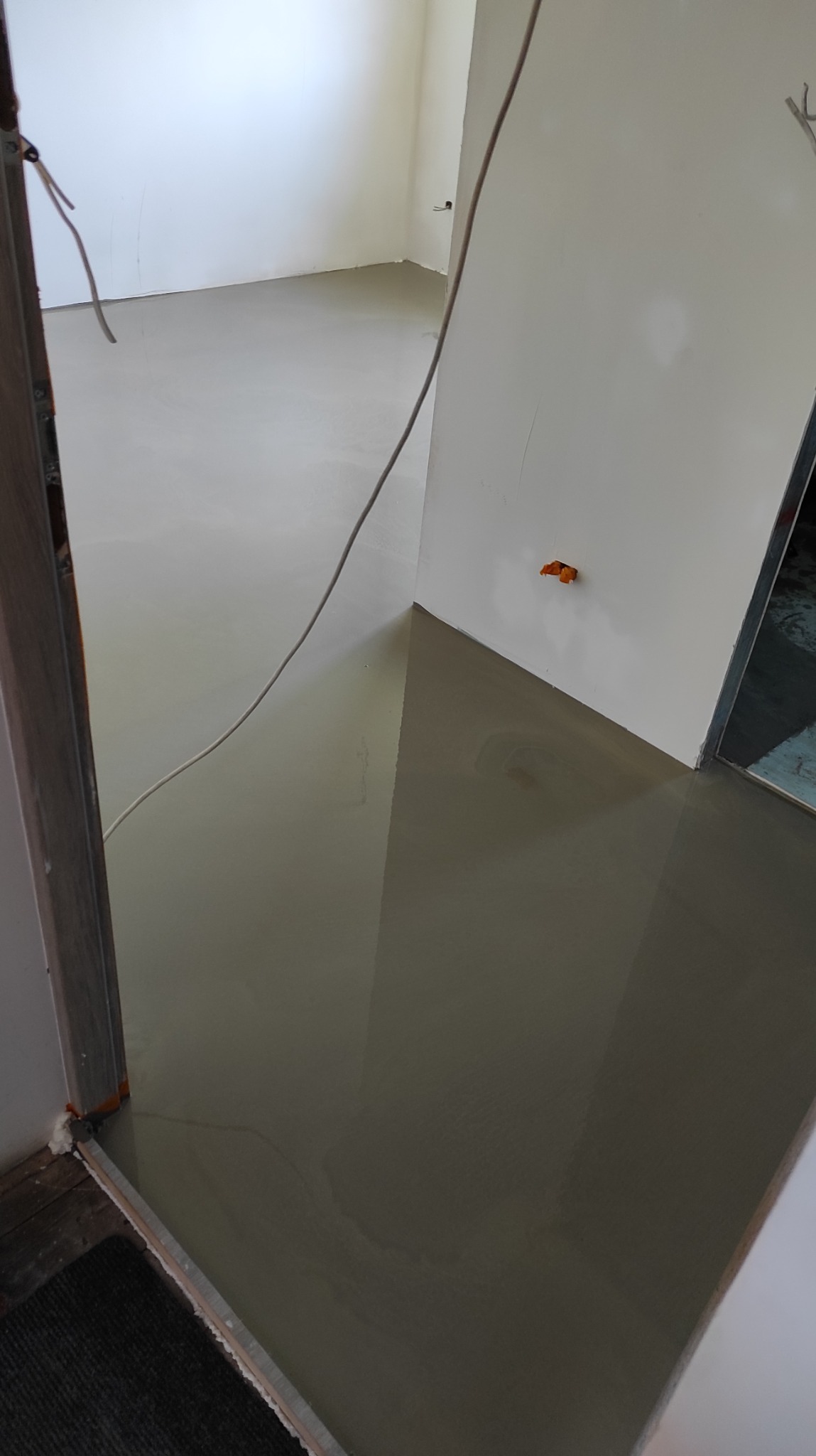 Wylany, mokry samopoziomujący podkład podłogowy w jasnym pomieszczeniu, widok z progu drzwi. Kabel elektryczny zwisa ze ściany.