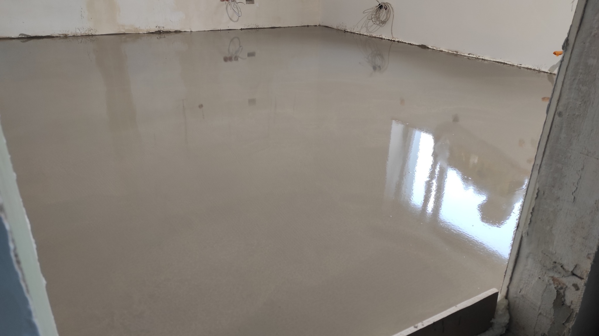 Wylana, gładka posadzka samopoziomująca w jasnym odcieniu szarości, odbijająca światło. Widoczne kable elektryczne wystające ze ściany. Surowy stan wykończenia.