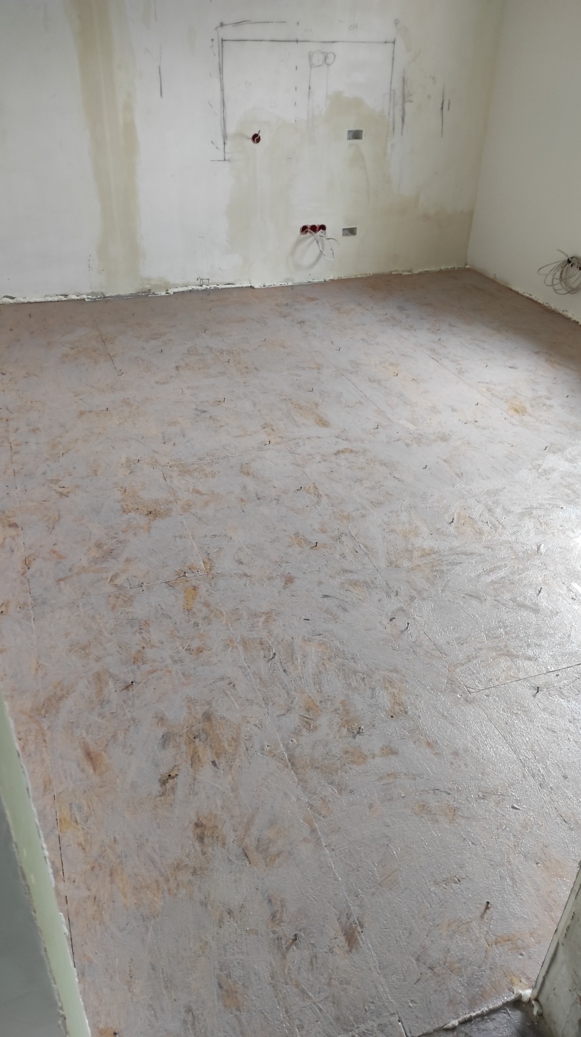 Przygotowane podłoże pod parkiet w jasnym pomieszczeniu z widocznymi śladami po szpachlowaniu ścian i instalacją elektryczną. Widok z góry.