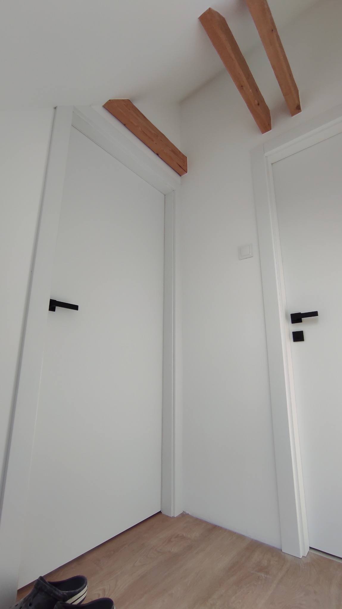 Białe drzwi z czarnymi klamkami w minimalistycznym wnętrzu. Widoczne drewniane belki na suficie i fragment podłogi. Ujęcie z dołu.