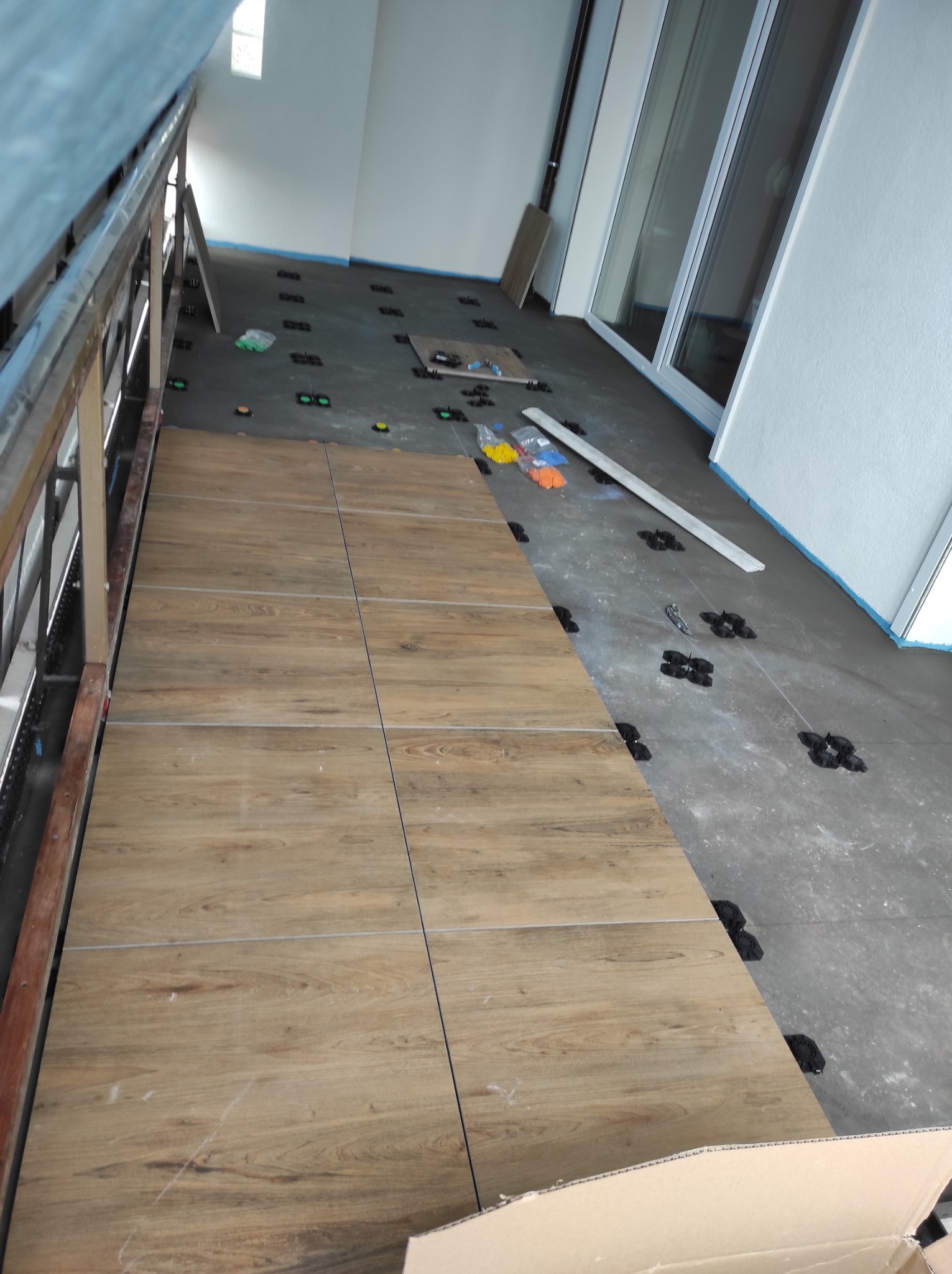 Układanie płytek imitujących drewno na schodach, widoczne systemy poziomowania, częściowo ułożone, z boku barierka, w tle okno. Prace wykończeniowe w trakcie.