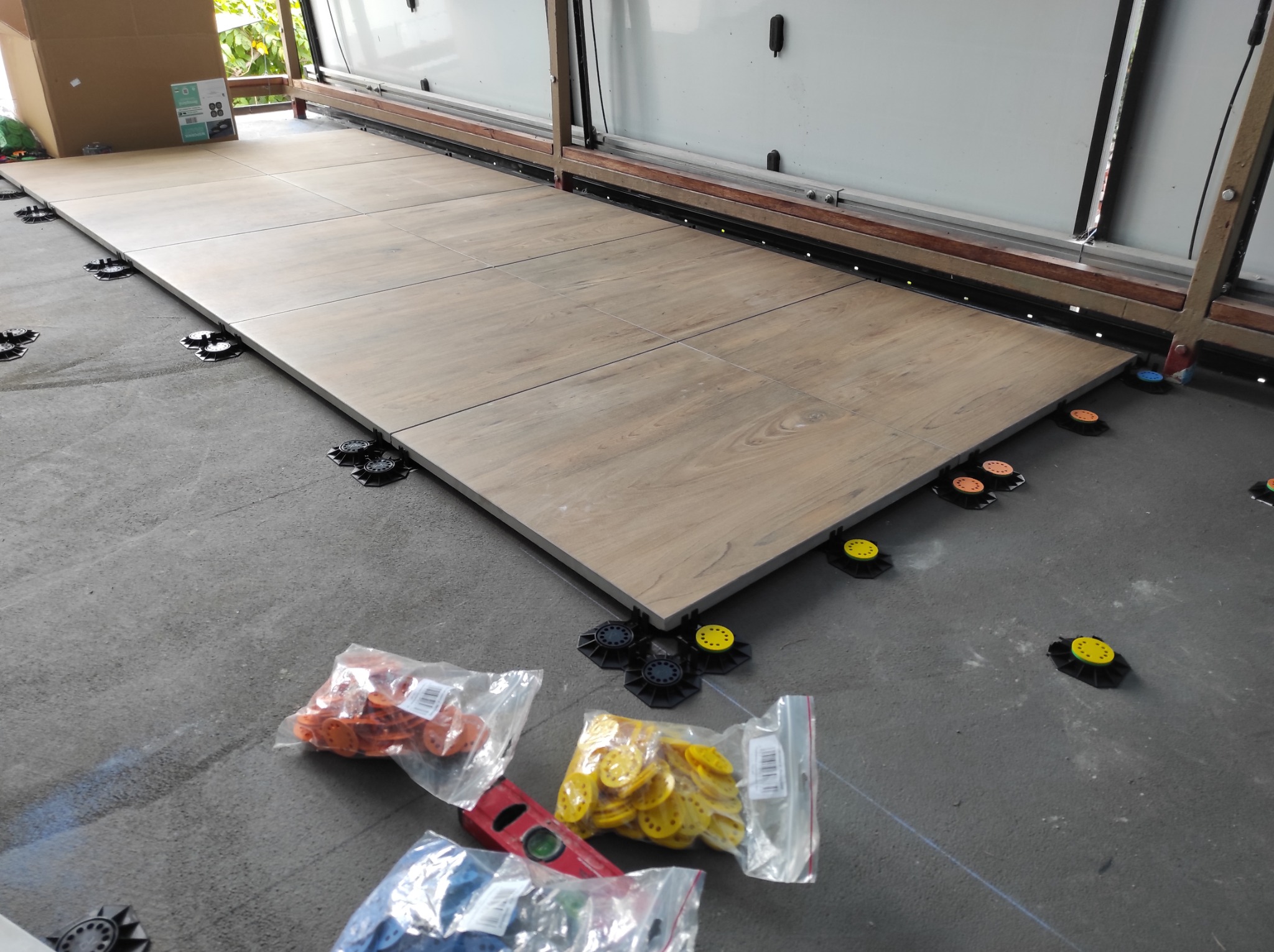 Układanie płytek podłogowych na tarasie z użyciem regulowanych wsporników. Widoczne poziomica i opakowania z elementami montażowymi.