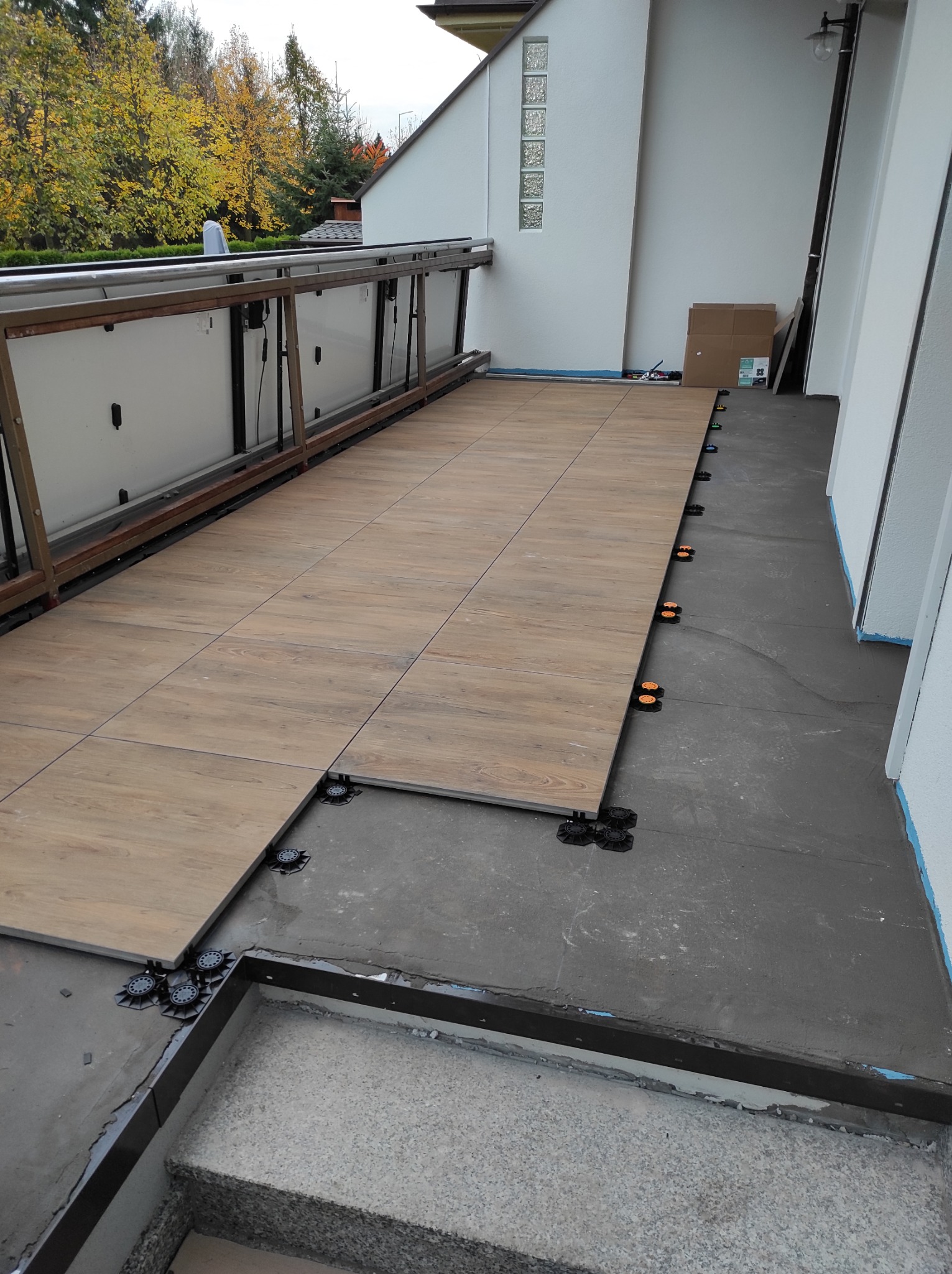 Montaż płytek imitujących drewno na balkonie z użyciem regulowanych wsporników. Widoczne fragmenty balustrady i schodów. Częściowo ułożona podłoga.