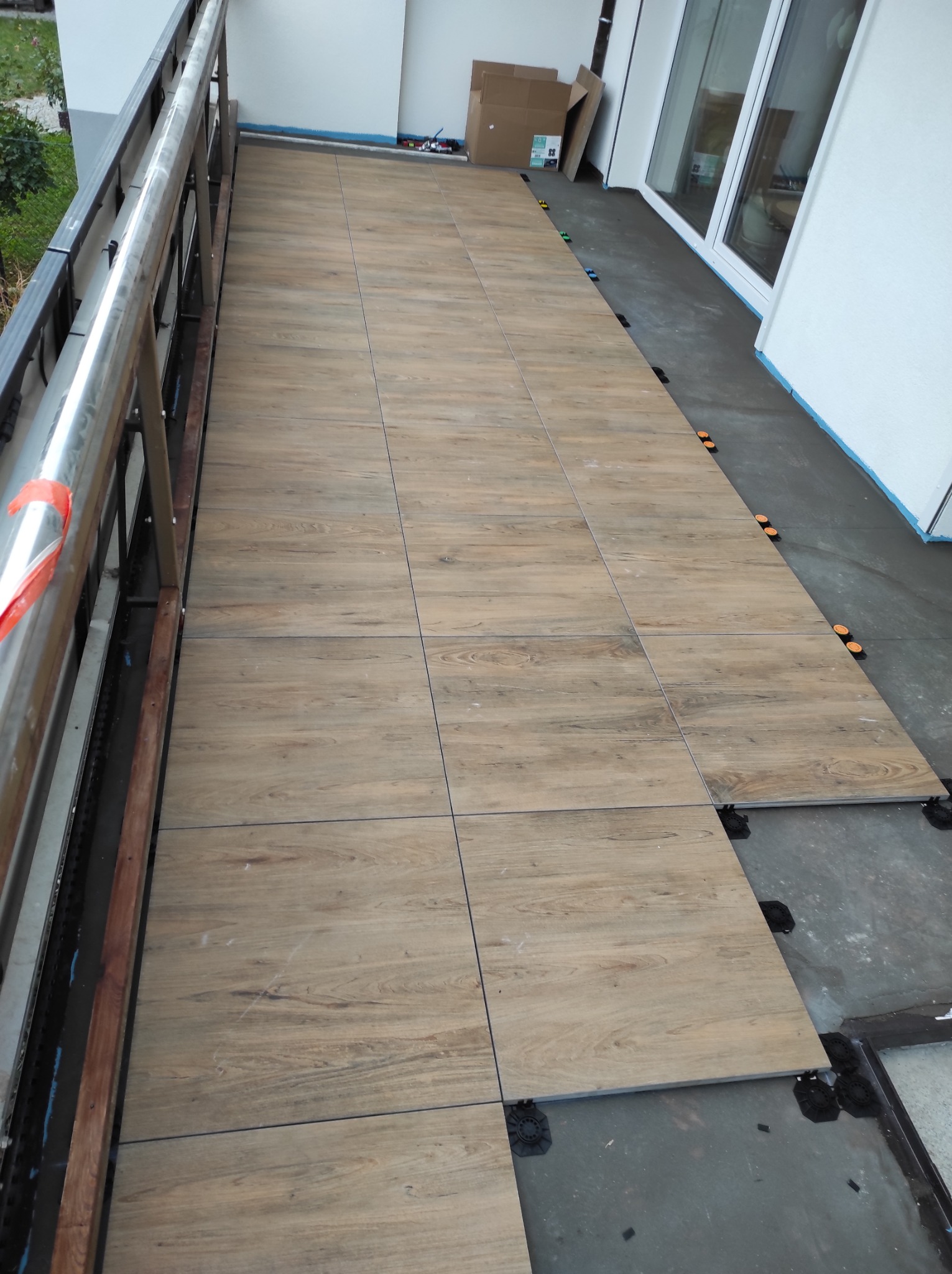 Układanie płytek imitujących drewno na balkonie, widoczne systemy poziomowania, balustrada po lewej, okna balkonowe w tle. Prace w toku.
