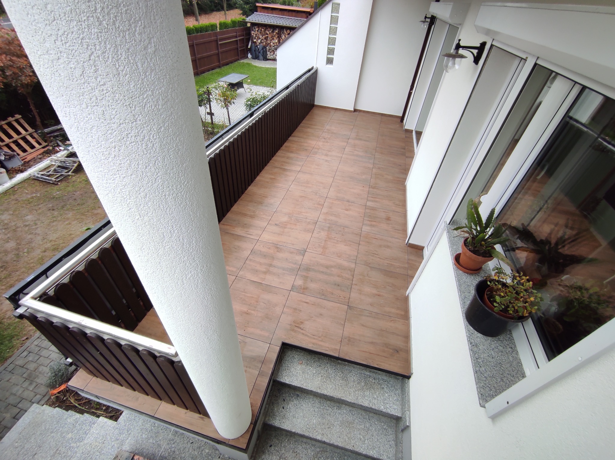 Wykończony balkon z brązowymi płytkami imitującymi drewno, białą kolumną i balustradą. Widok z góry na schody wejściowe i otoczenie domu.