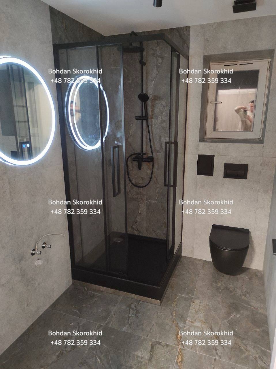 Nowoczesna łazienka z czarną kabiną prysznicową, okrągłym lustrem z podświetleniem LED i wiszącą toaletą. Szare płytki imitujące marmur na ścianach i podłodze.