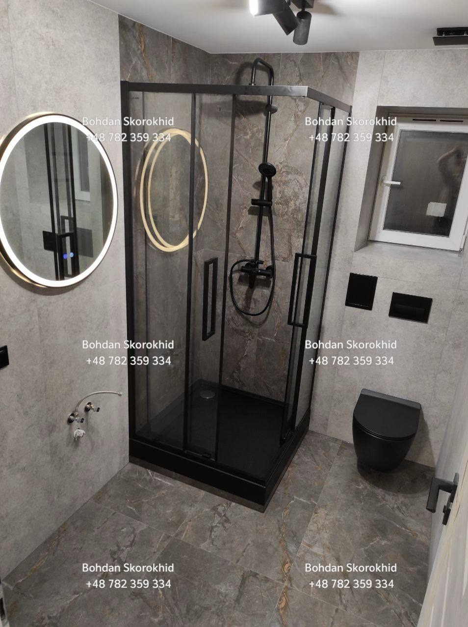 Nowoczesna łazienka z czarną kabiną prysznicową, okrągłym lustrem z podświetleniem LED i szarą marmurową płytką. Minimalistyczny design, eleganckie wykończenie.