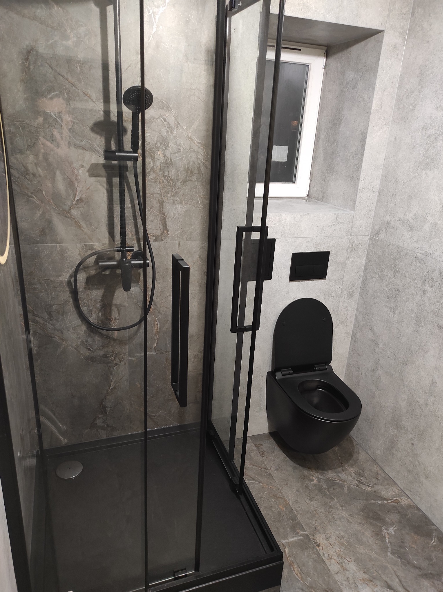 Nowoczesna łazienka w szarościach z czarnymi akcentami: kabina prysznicowa typu walk-in, podwieszana toaleta i okno. Minimalistyczny design, płytki imitujące marmur.