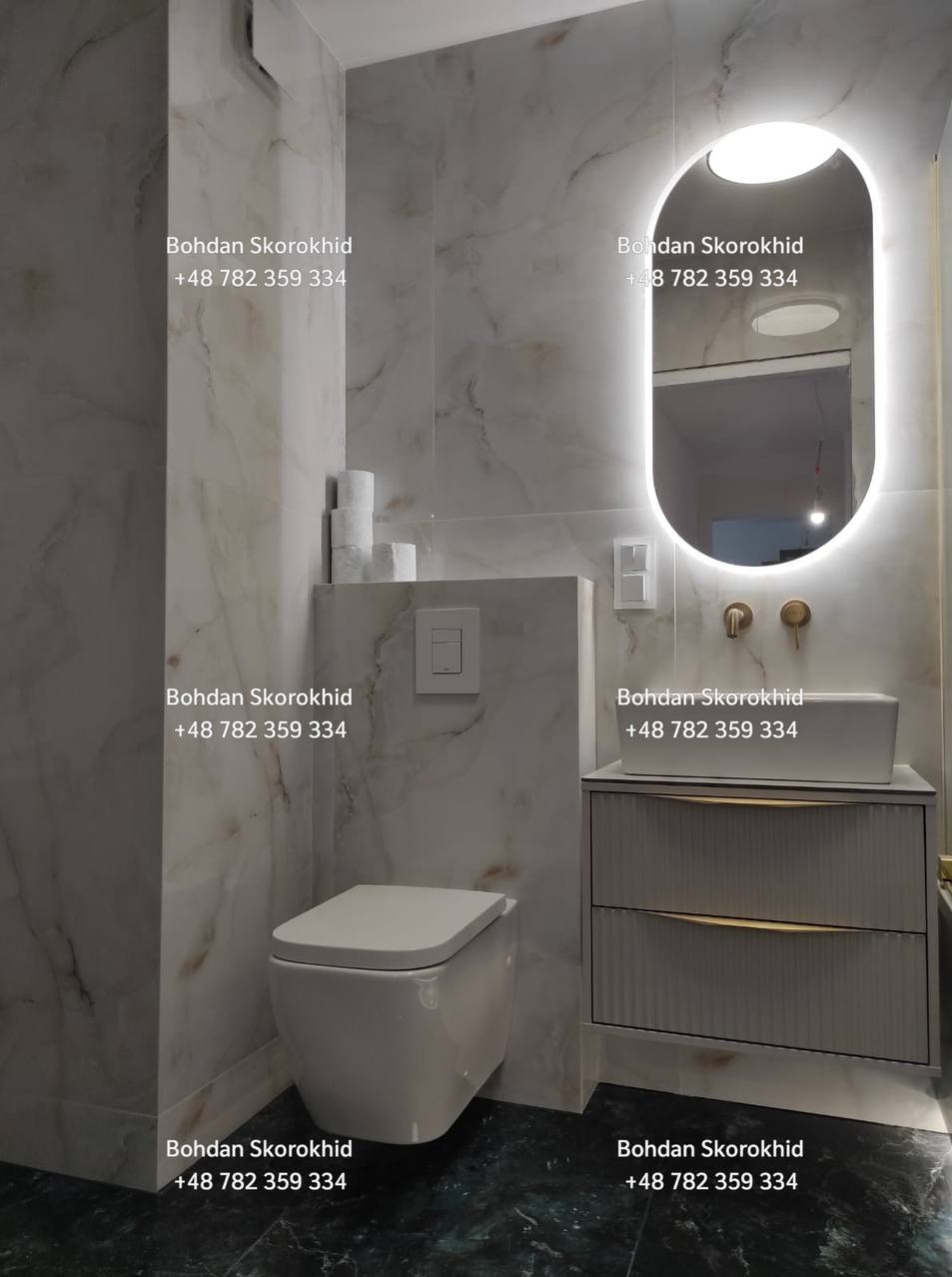 Elegancka łazienka z marmurowymi płytkami, podwieszaną toaletą i szafką z umywalką podświetloną owalnym lustrem LED. Nowoczesny design, złote akcenty.