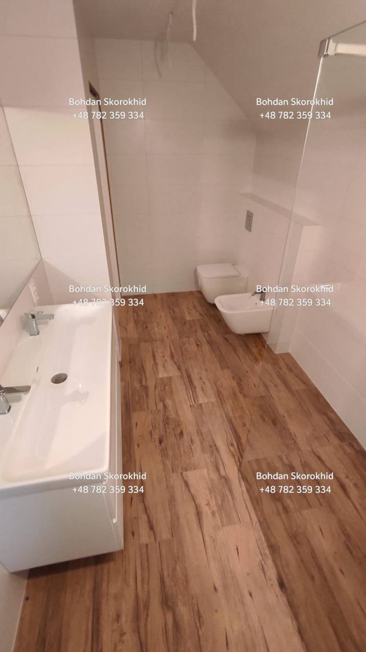 Jasna łazienka po remoncie z drewnopodobną podłogą, białą armaturą i minimalistycznym designem. Widoczne umywalki, toaleta i kabina prysznicowa.