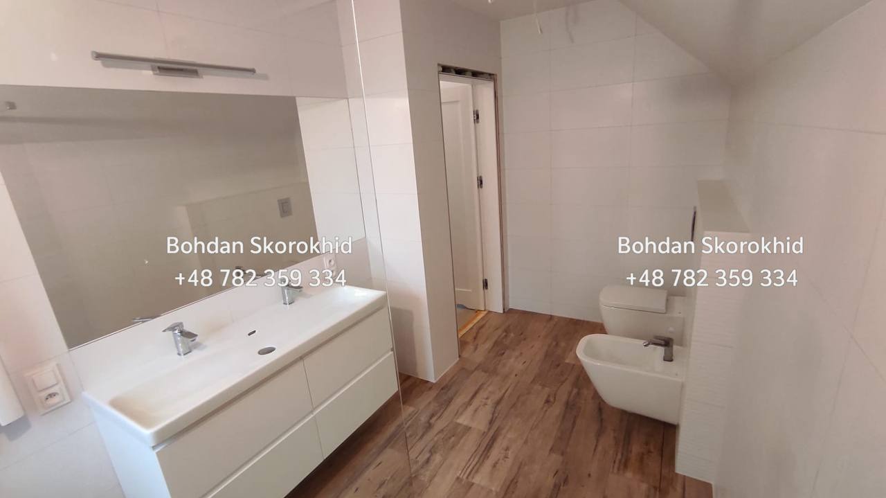 Jasna łazienka po remoncie: białe płytki, drewnopodobna podłoga, umywalka z szafką i duże lustro. Minimalistyczny design z widocznym fragmentem toalety i bidetu.