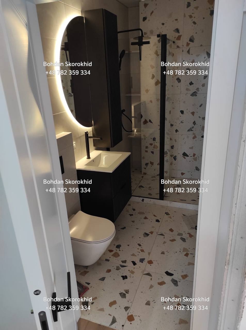 Nowoczesna łazienka z kabiną prysznicową typu walk-in, umywalką z szafką w kolorze czarnym i okrągłym lustrem z podświetleniem LED. Na podłodze i ścianach płytki lastryko.