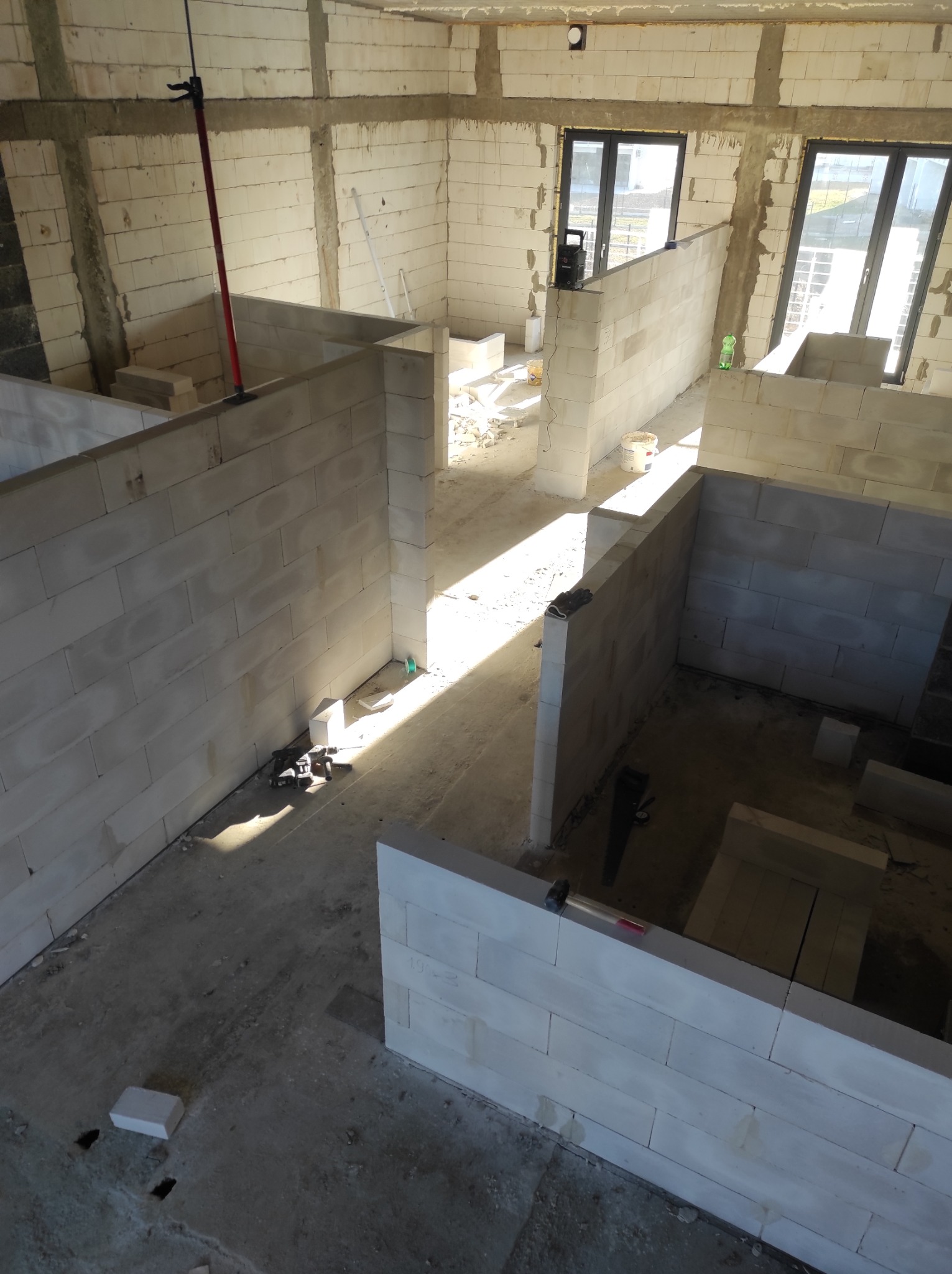 Wnętrze w trakcie budowy z białymi ścianami z betonu komórkowego, widoczne okna i narzędzia, surowy stan, jasne światło dzienne.