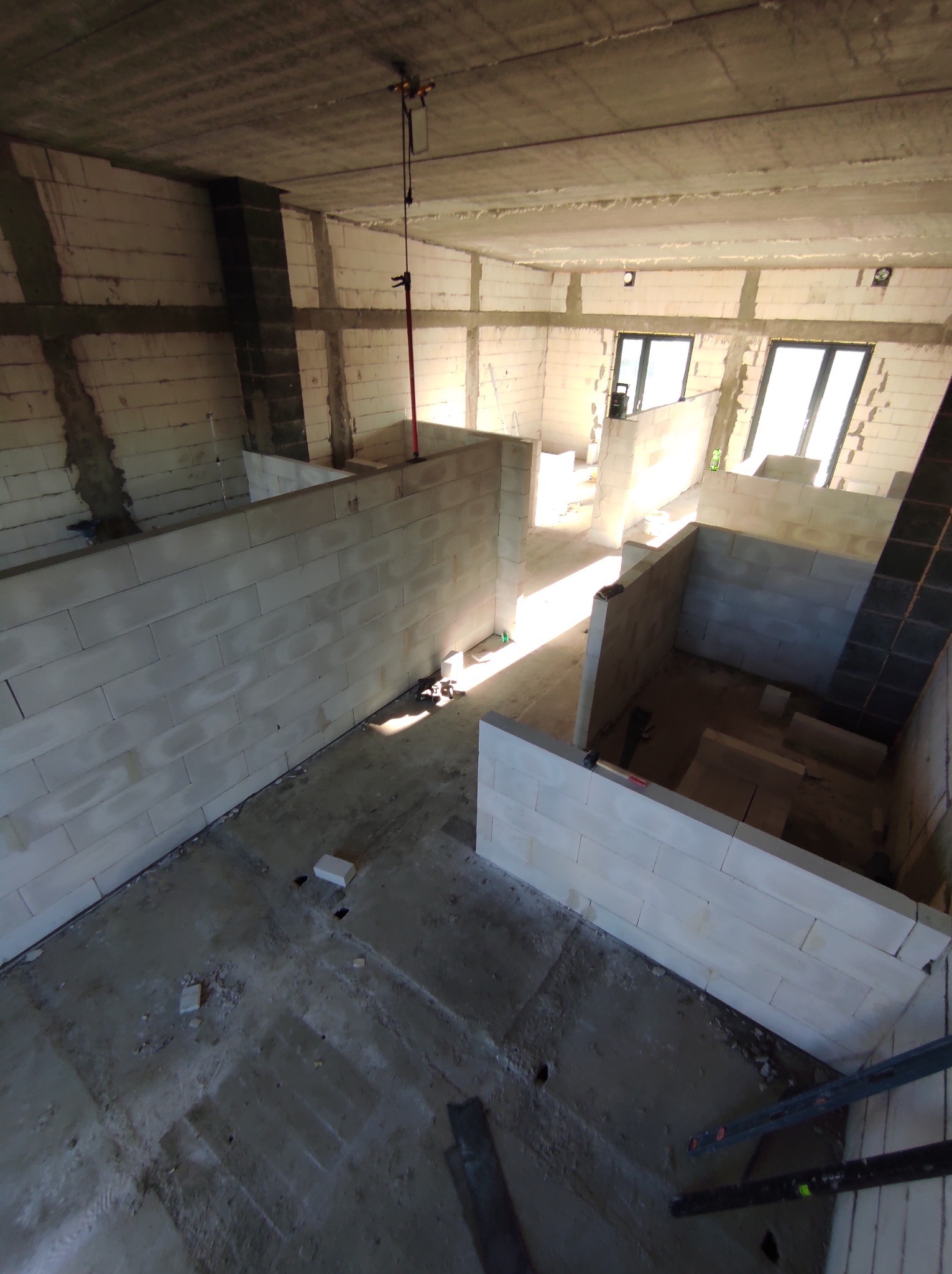 Wnętrze w trakcie budowy z białymi ścianami z bloczków, widoczne okna i surowy betonowy strop. Na podłodze resztki materiałów budowlanych i narzędzia.